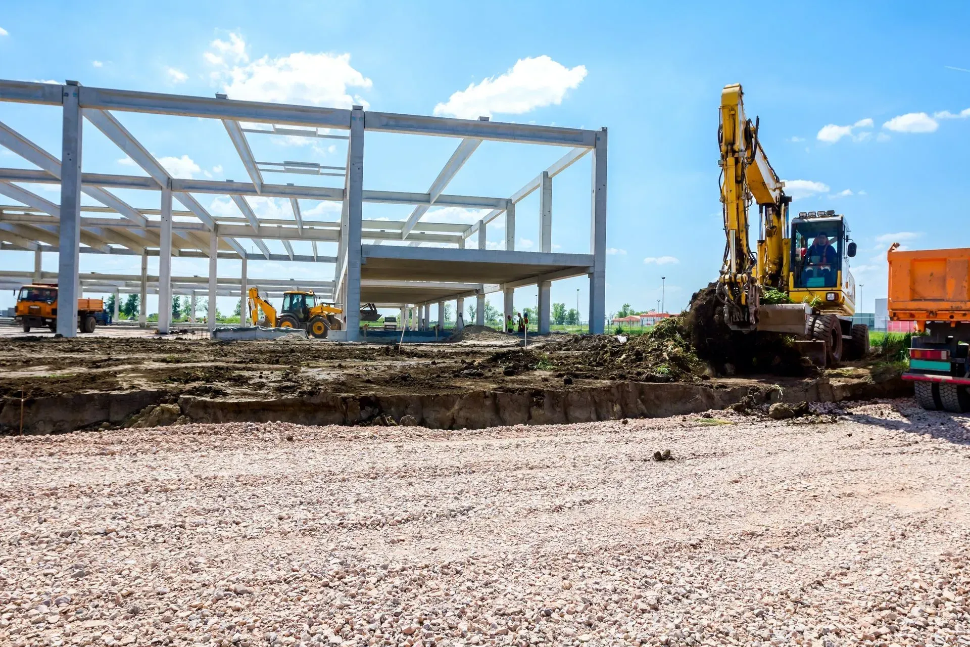 Construction site with steel building frame and yellow excavator on gravel under a blue sky