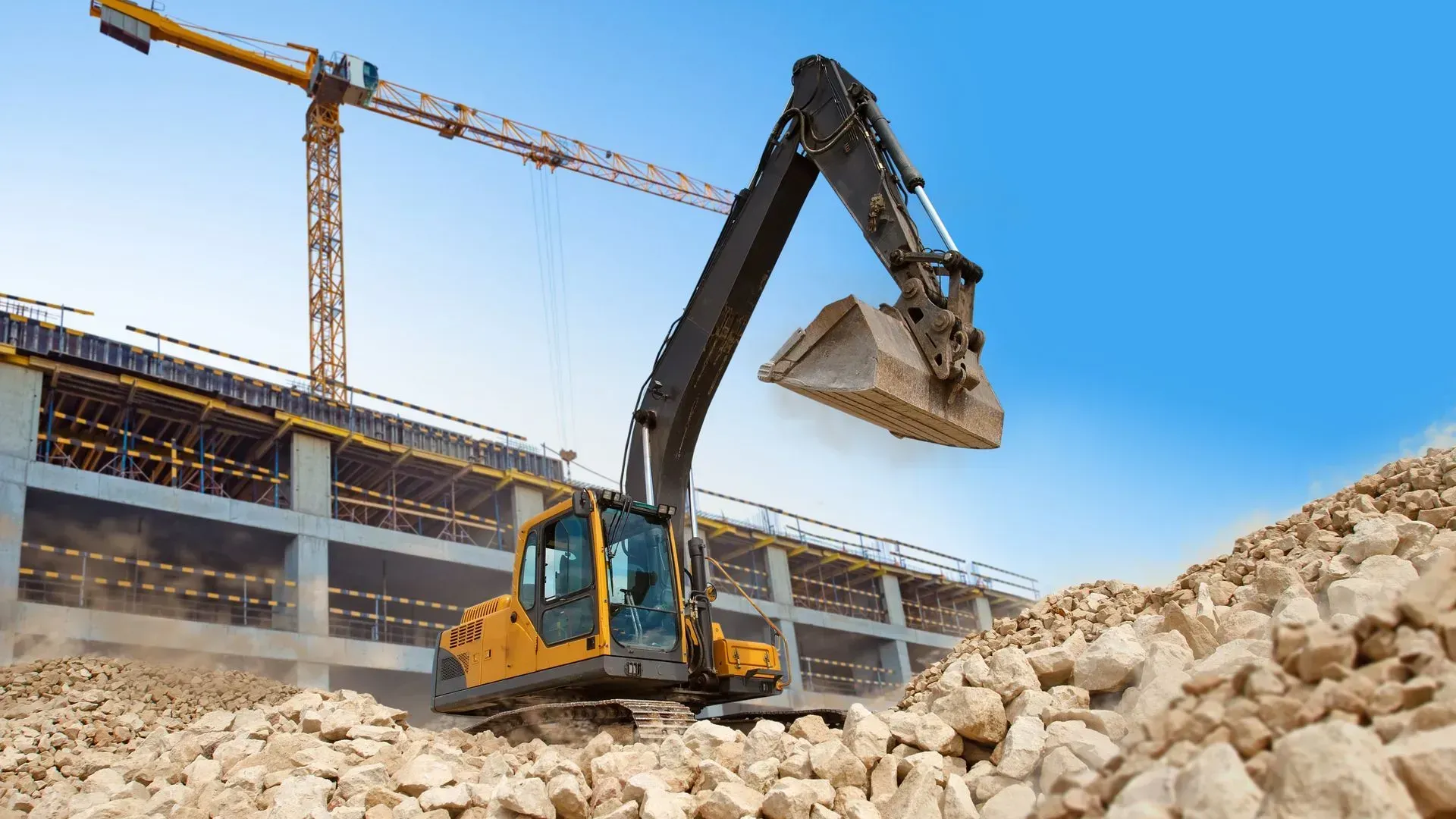 Excavator loading rocks at a construction site with cranes and a partially built building in the background