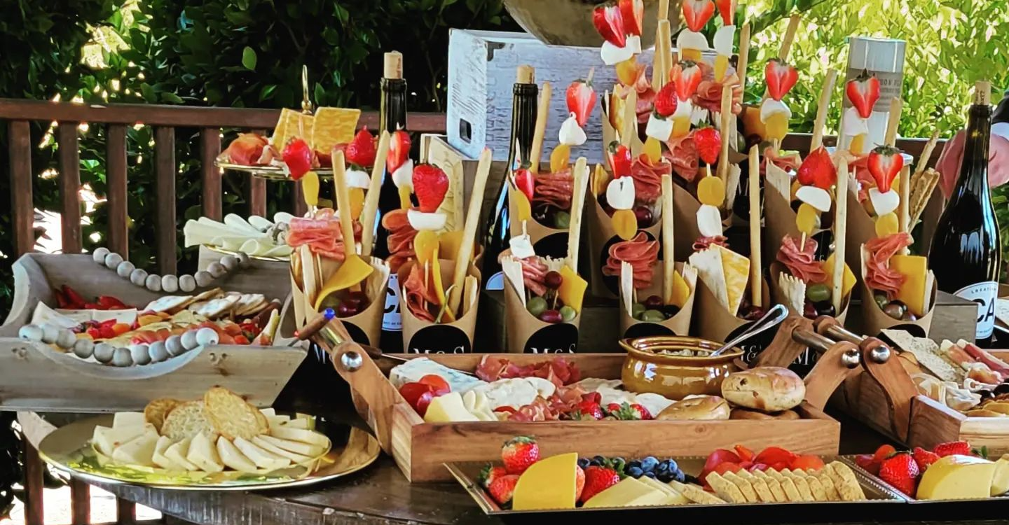 Charcuterie spread with various cheeses, meats, crackers, and fruit skewers on a wooden table outdoors.