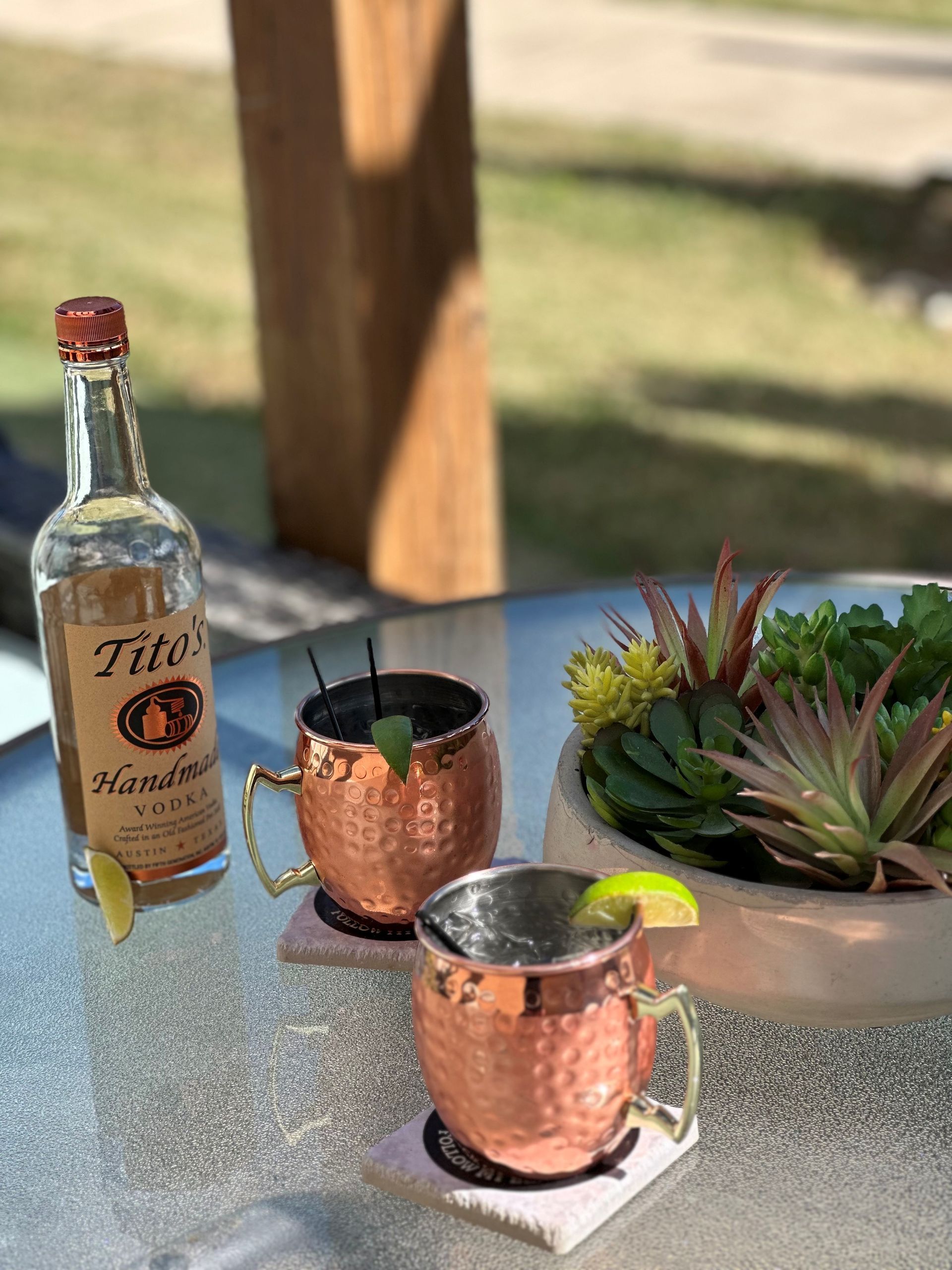 Two copper mugs, a Tito's vodka bottle, lime wedges, and a succulent planter sit on a glass table outdoors.