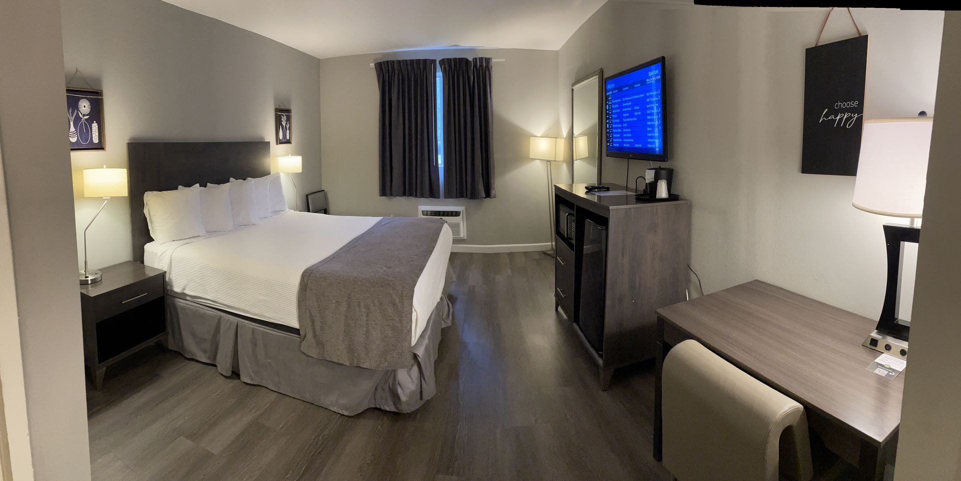 Hotel room with a bed, desk, and TV. Gray walls, bed skirt, and furniture. Dark curtains and wood floors.