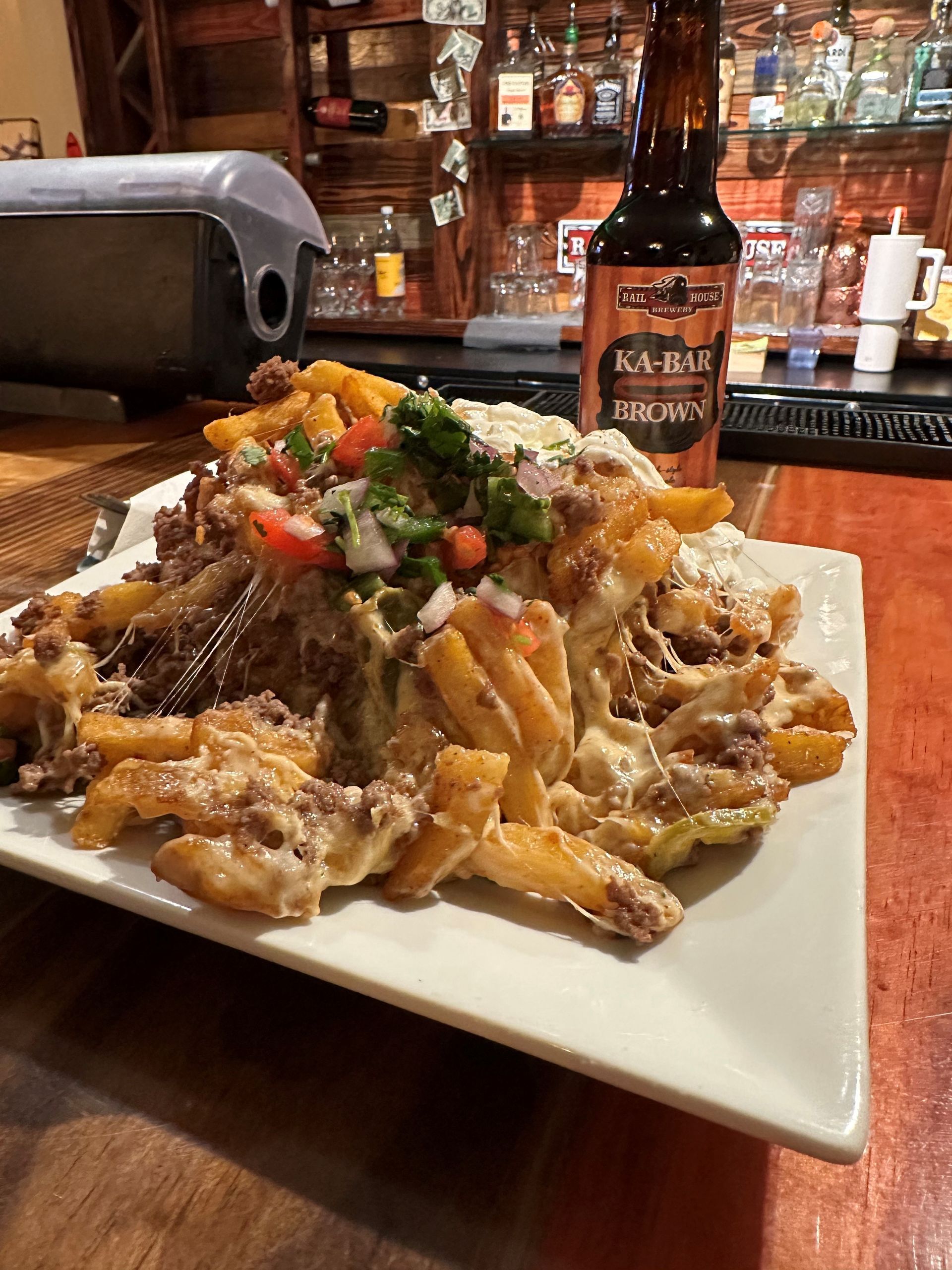 Loaded fries with meat and toppings on a white plate, with a beer bottle in the background at a bar.