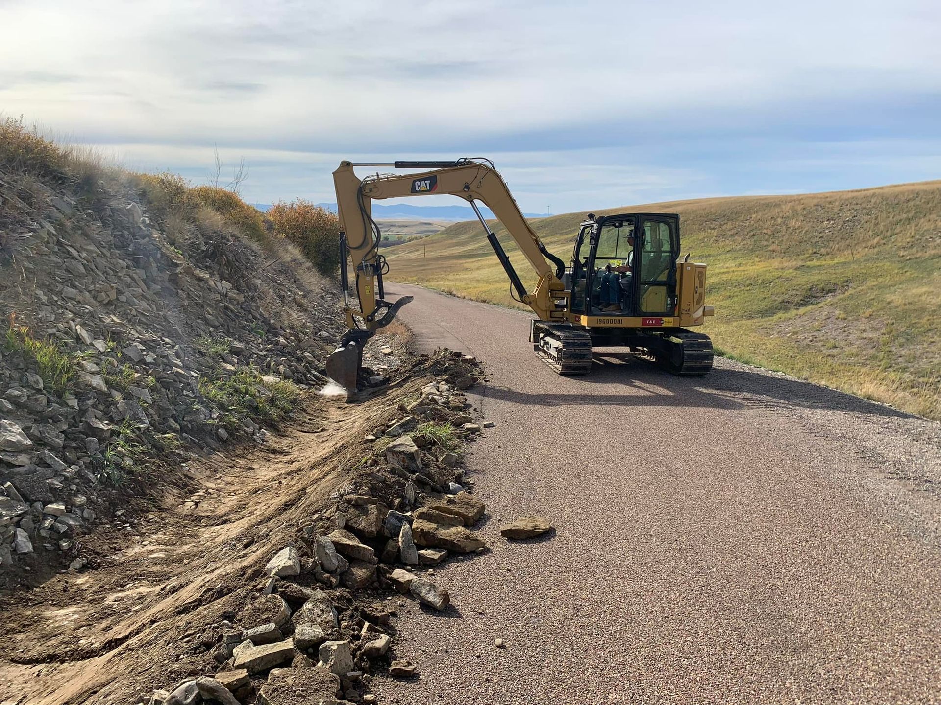 Excavator working along a gravel road between grassy hills, digging a trench on the left side.