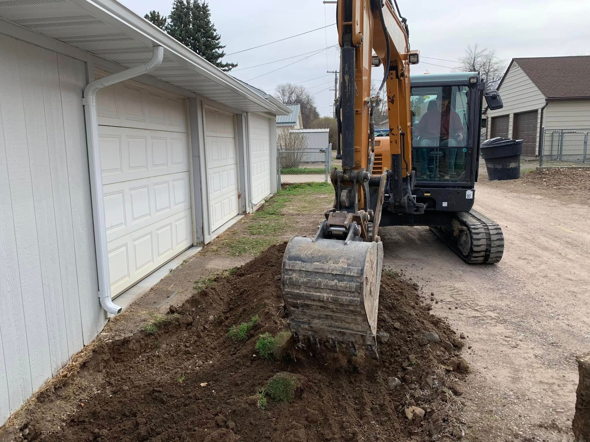 Excavator clearing soil beside a white garage in a residential driveway.
