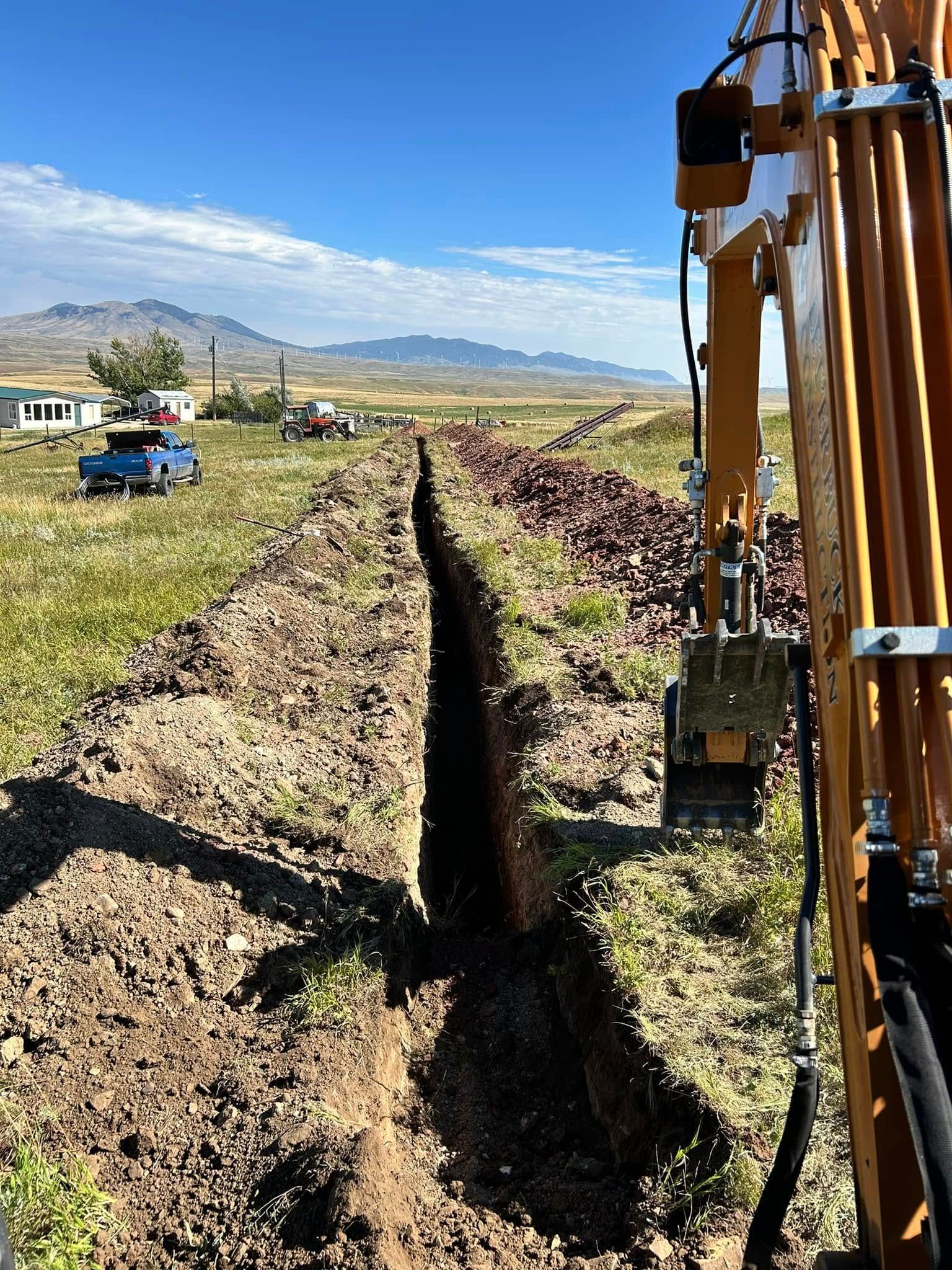 Excavator digging a long trench in a rural field under a blue sky with mountains in the distance
