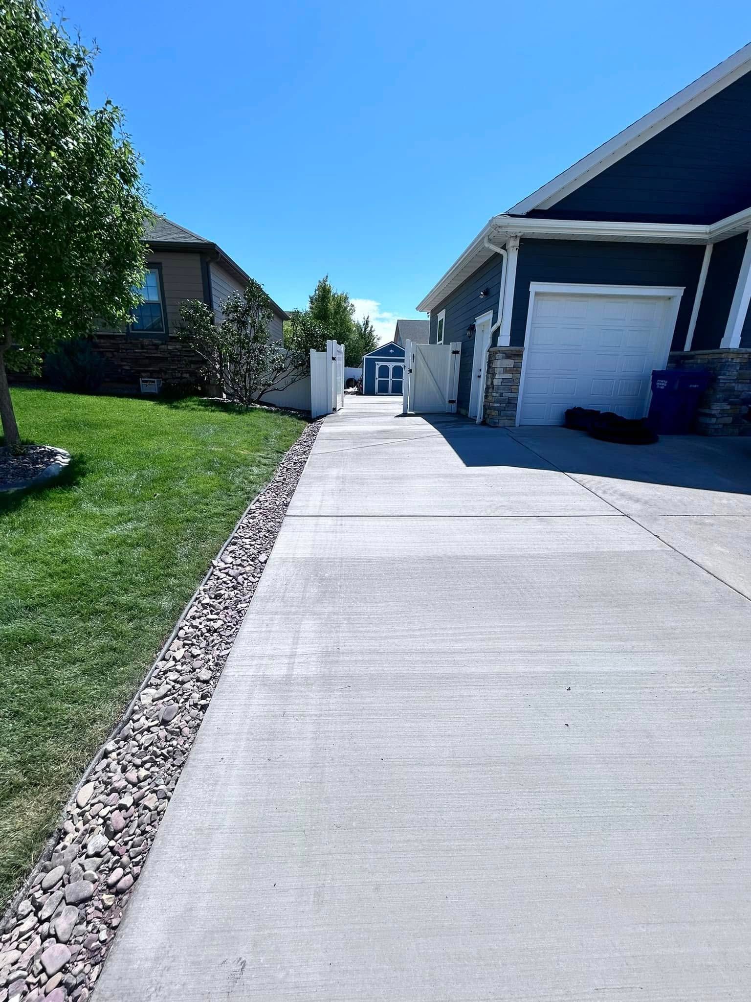 Long concrete driveway between houses under a clear blue sky, leading to a closed gate.