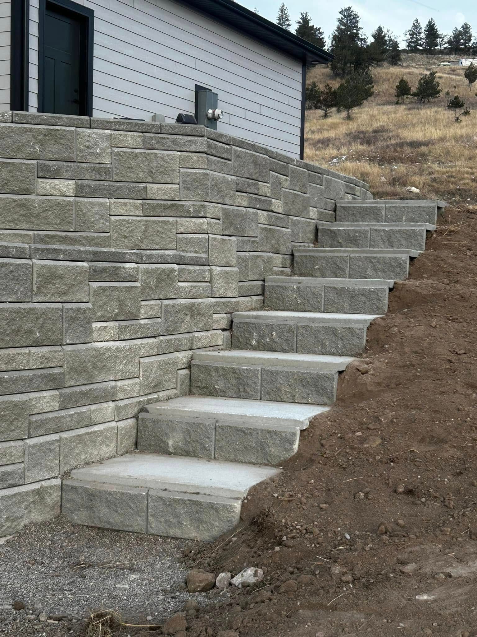 Stone retaining wall with concrete steps leading up a dirt slope beside a house.