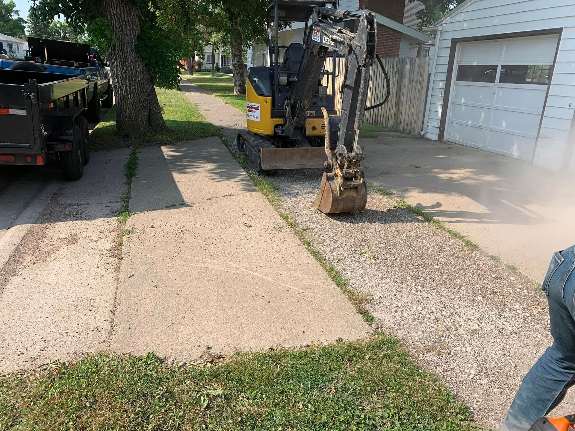 Excavator parked beside a driveway and garage, with gravel being spread over a path and a truck nearby
