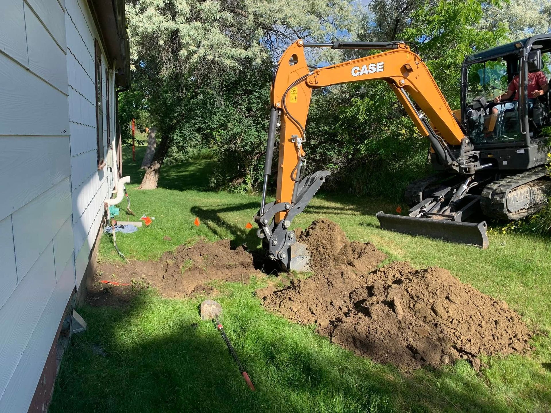 Orange excavator digging a trench beside a blue house, with a dirt pile in a grassy yard.