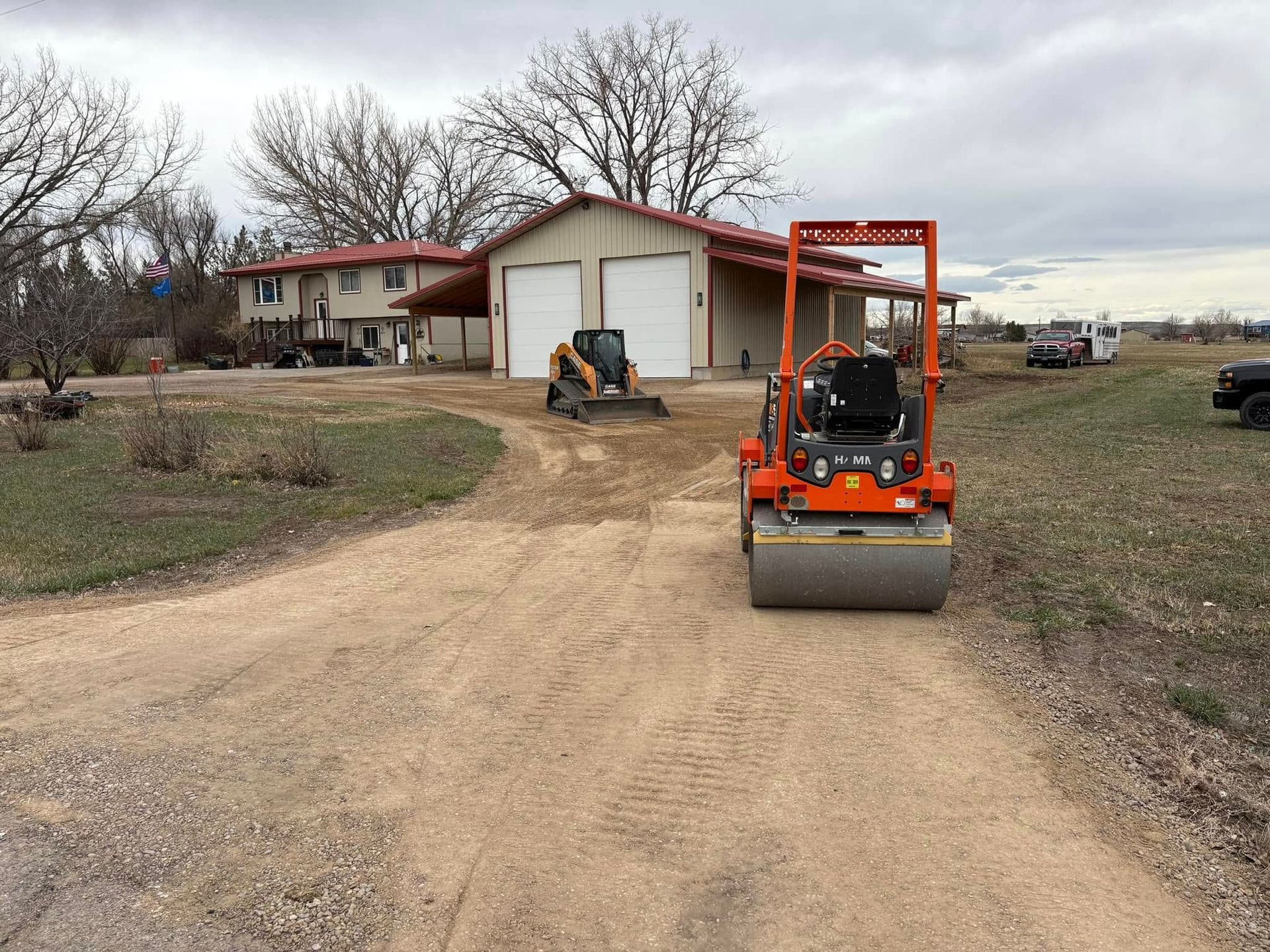Orange road roller on a gravel driveway in front of a house and garage, with a skid steer nearby