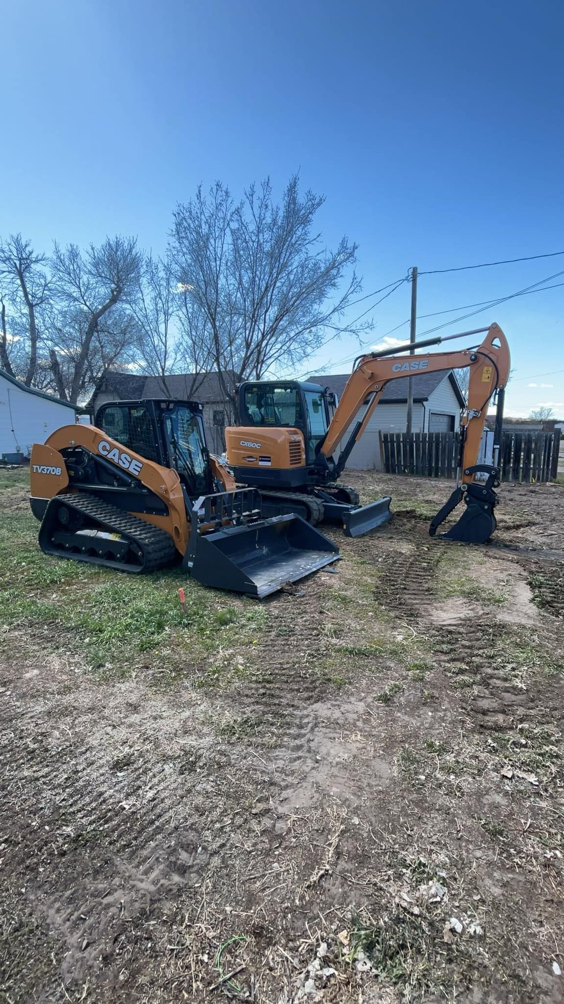 Orange excavator and compact loader parked on gravel near trees under a clear blue sky.