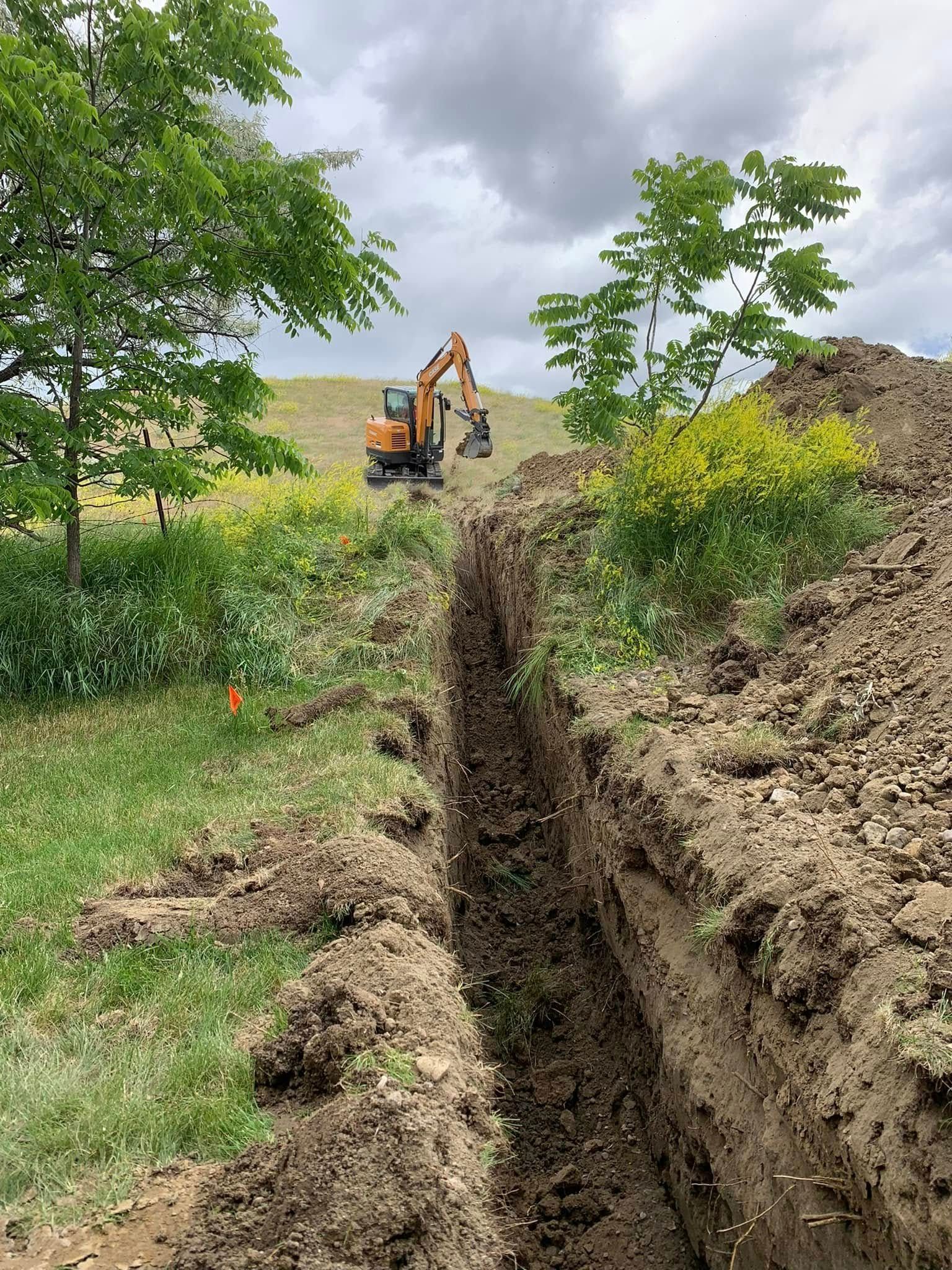 Excavator digging a deep trench beside a grassy field under cloudy skies.