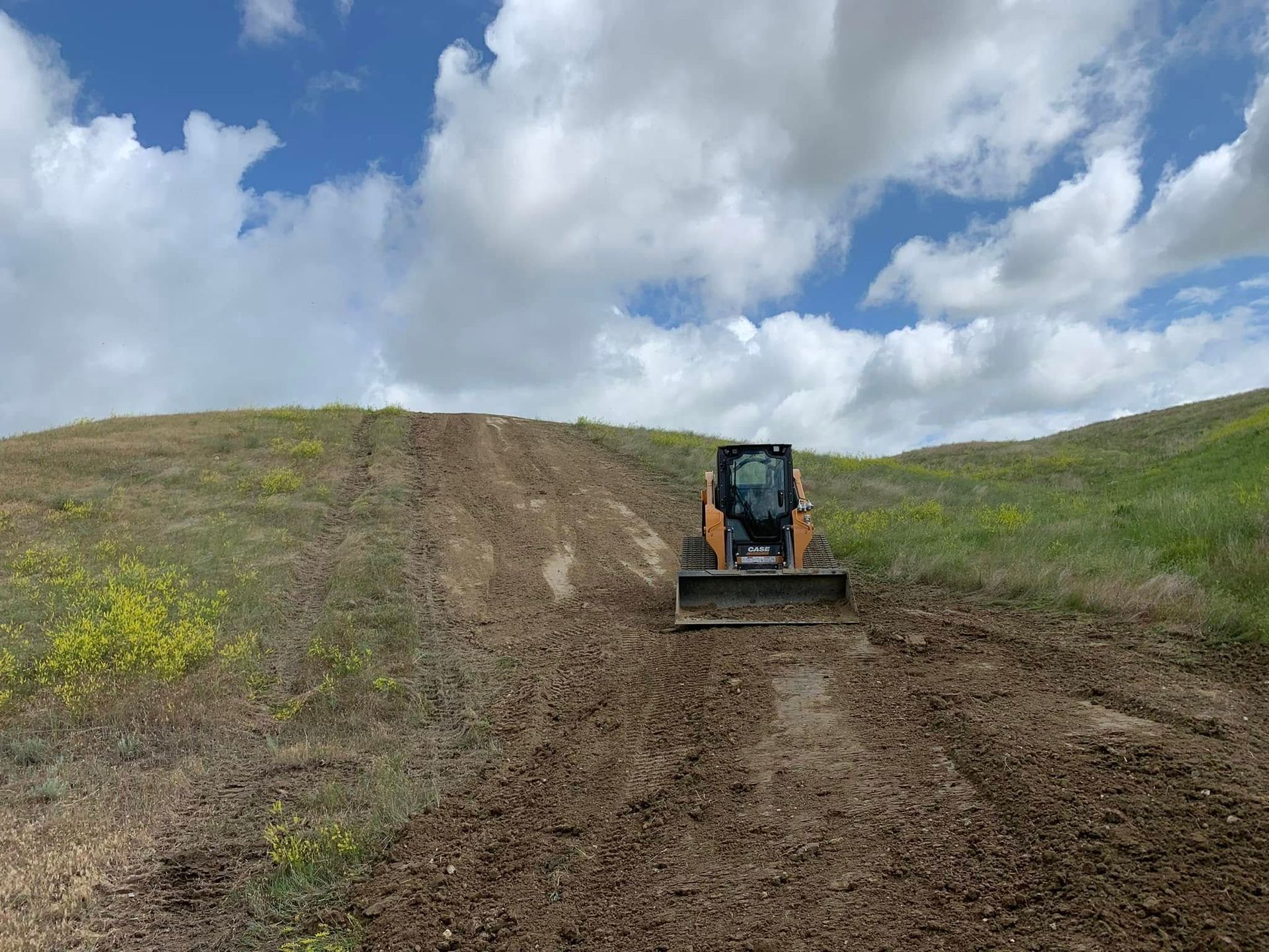 Bulldozer clearing a dirt path on a grassy hill under a cloudy sky