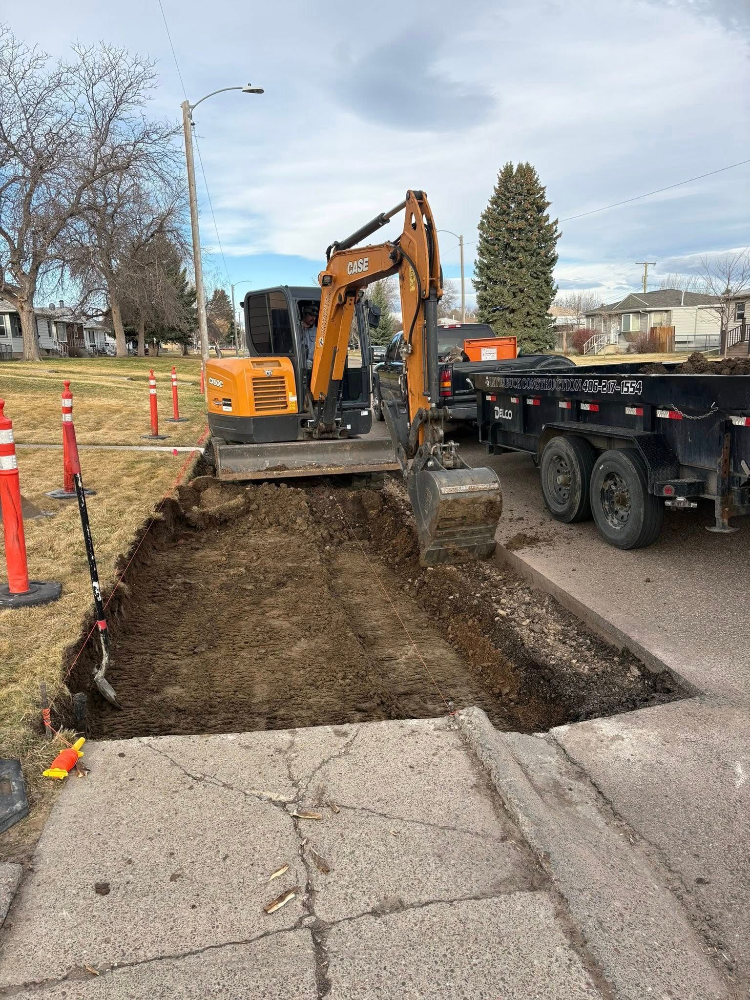 Excavator digging trench beside a road, with orange cones and a trailer on a construction site