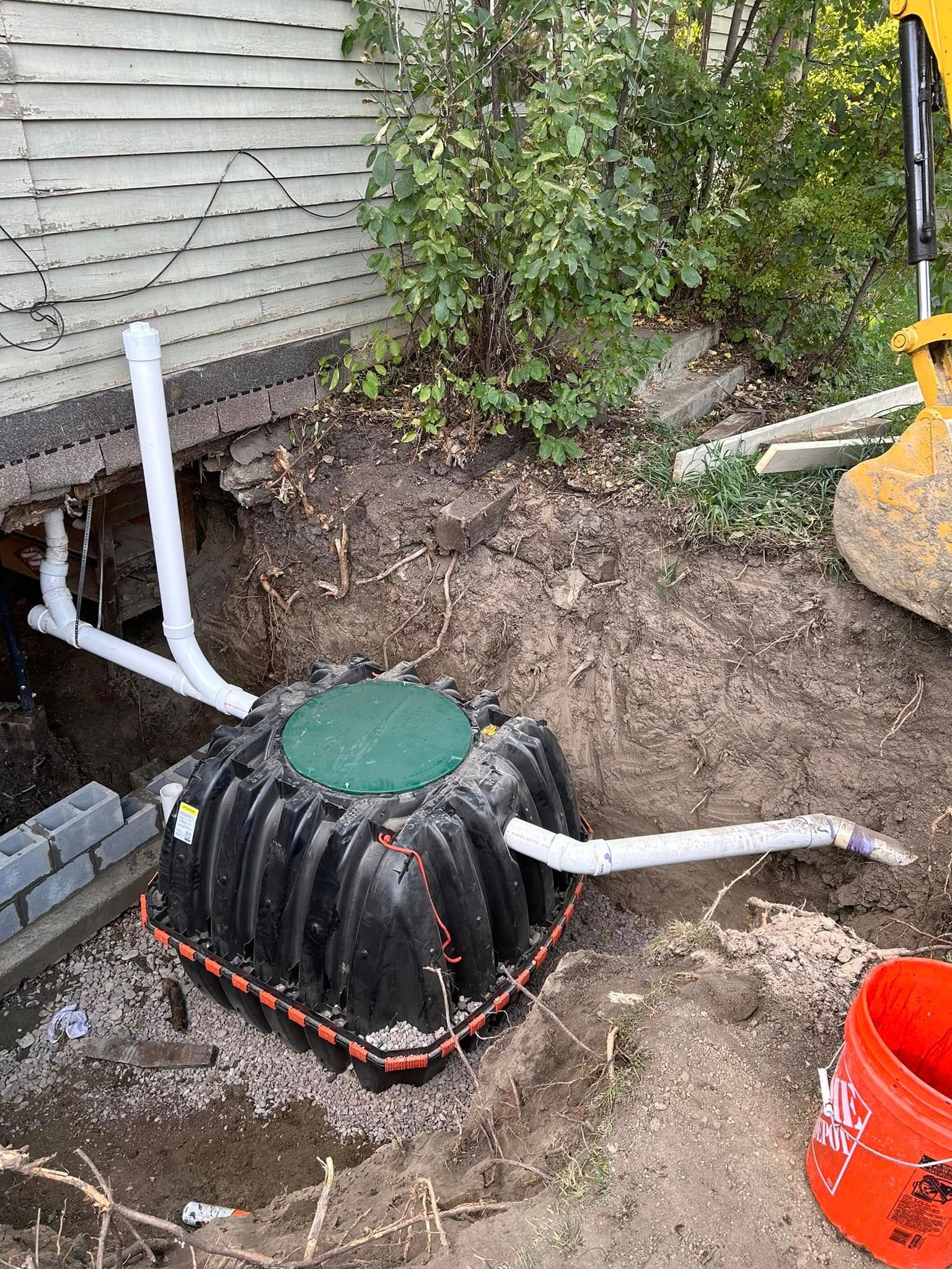 Backyard septic tank with green access cover, white pipes, and exposed dirt beside a house foundation
