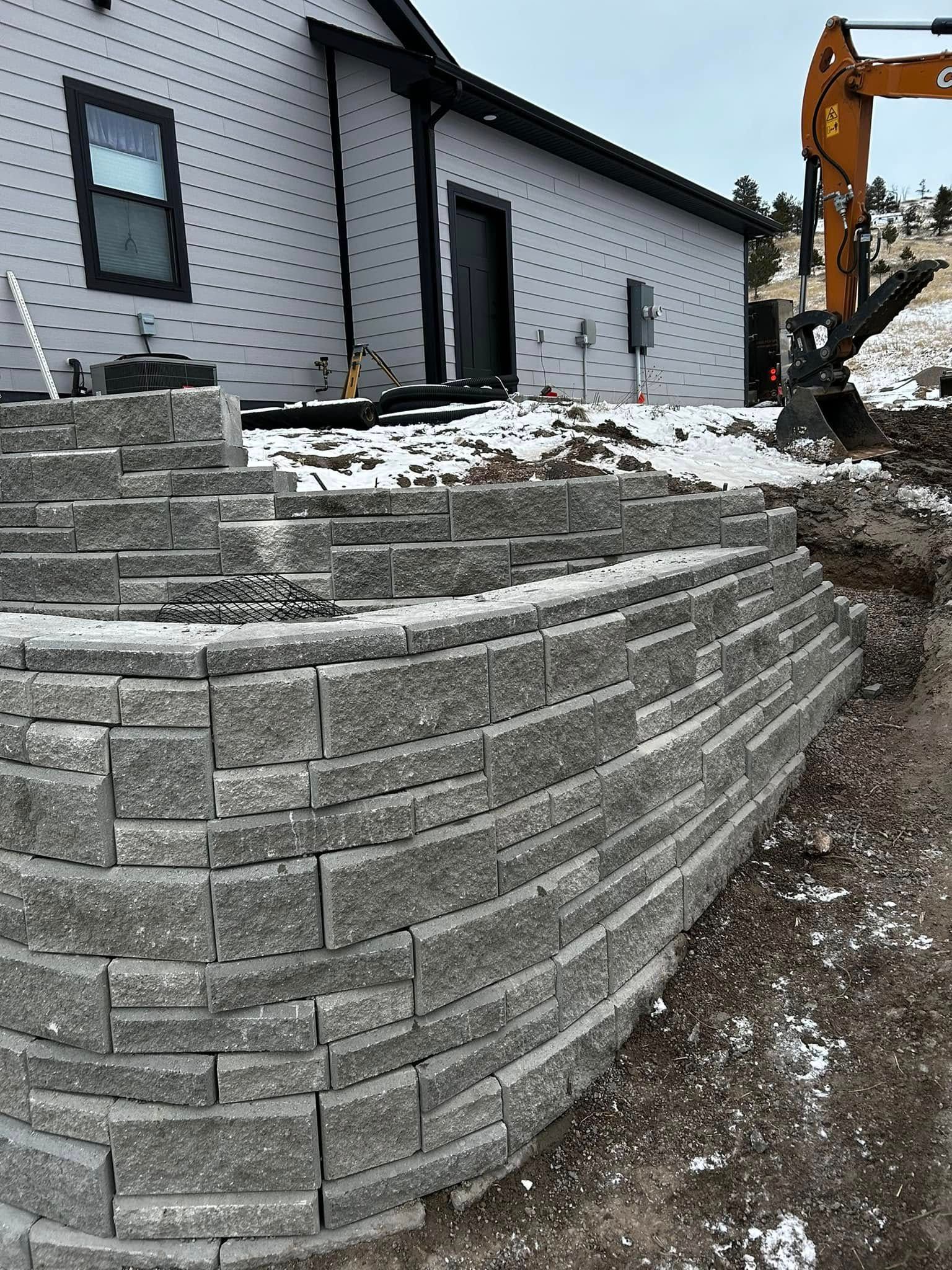 Retaining wall of stacked gray stone beside a white house, with an excavator and muddy construction site.