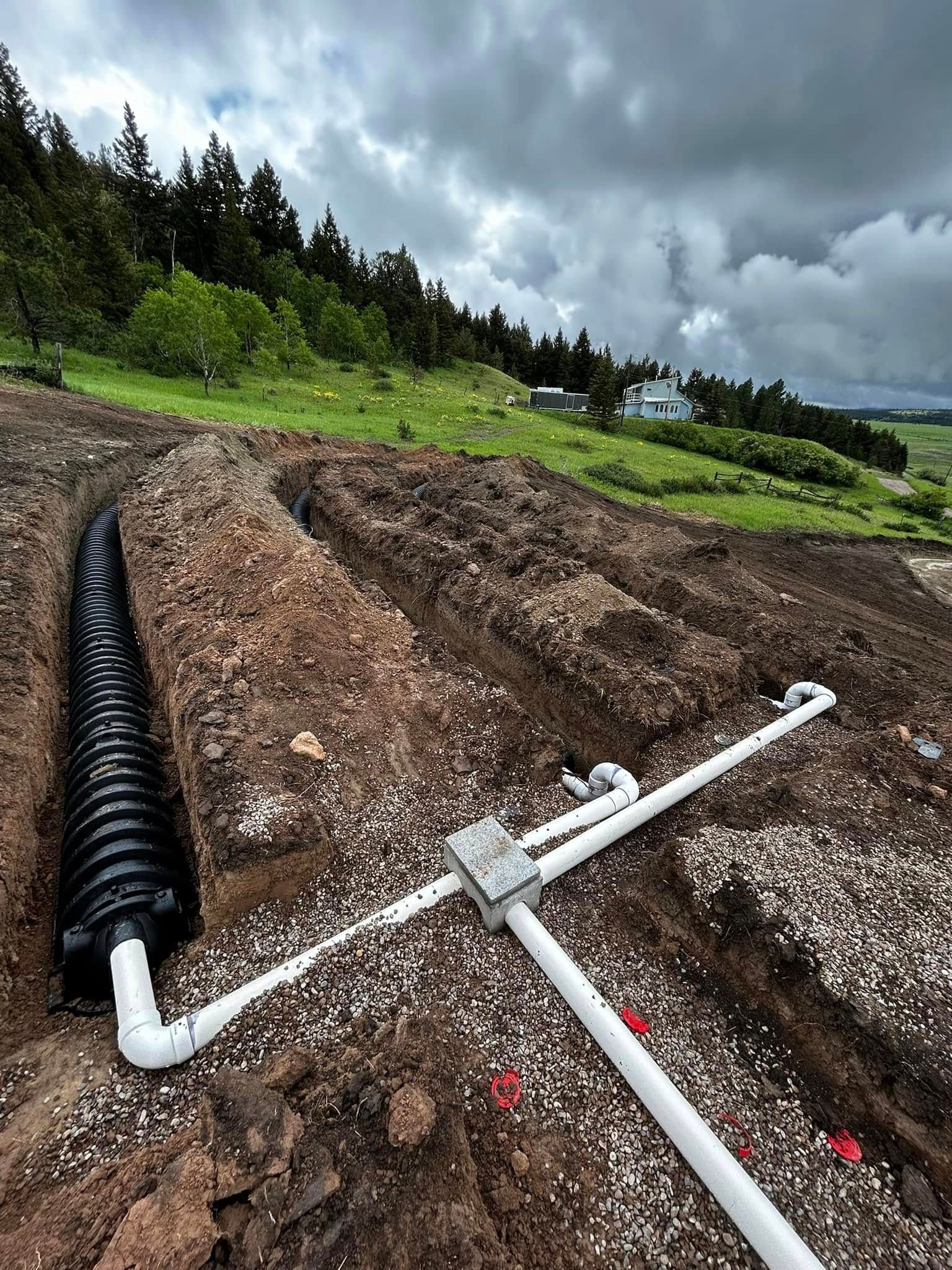 White drainage pipes installed in a dirt trench on a grassy hillside under cloudy skies