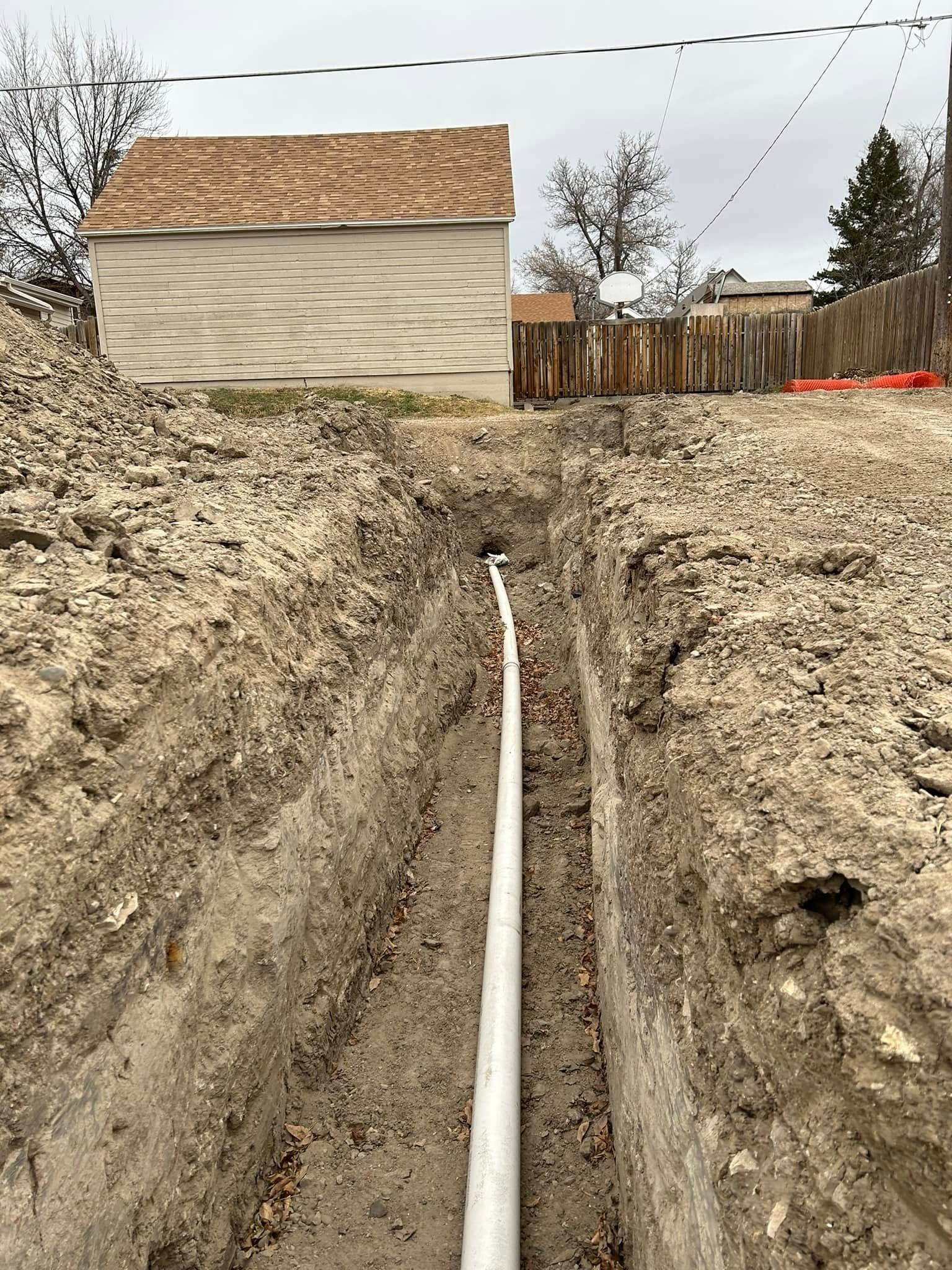 Trenched yard with a white utility pipe running toward a house and wooden fence.