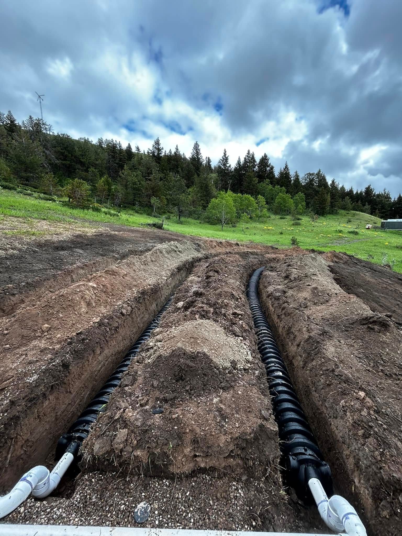 Freshly dug garden beds with black irrigation tubes under a cloudy sky and trees in the background