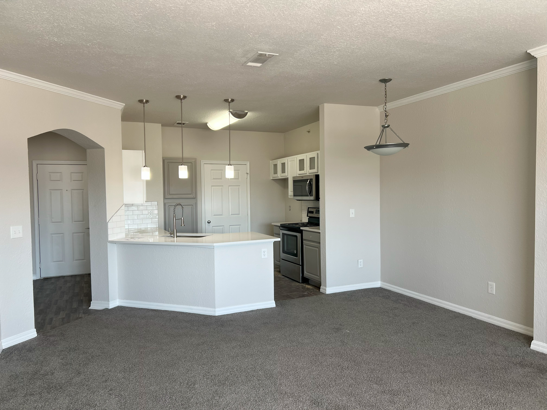 Empty carpeted living room opening to a kitchen with white cabinets and pendant lights.