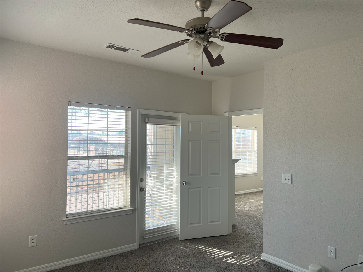 Empty bedroom with carpet, white walls, ceiling fan, and open door beside two windows.