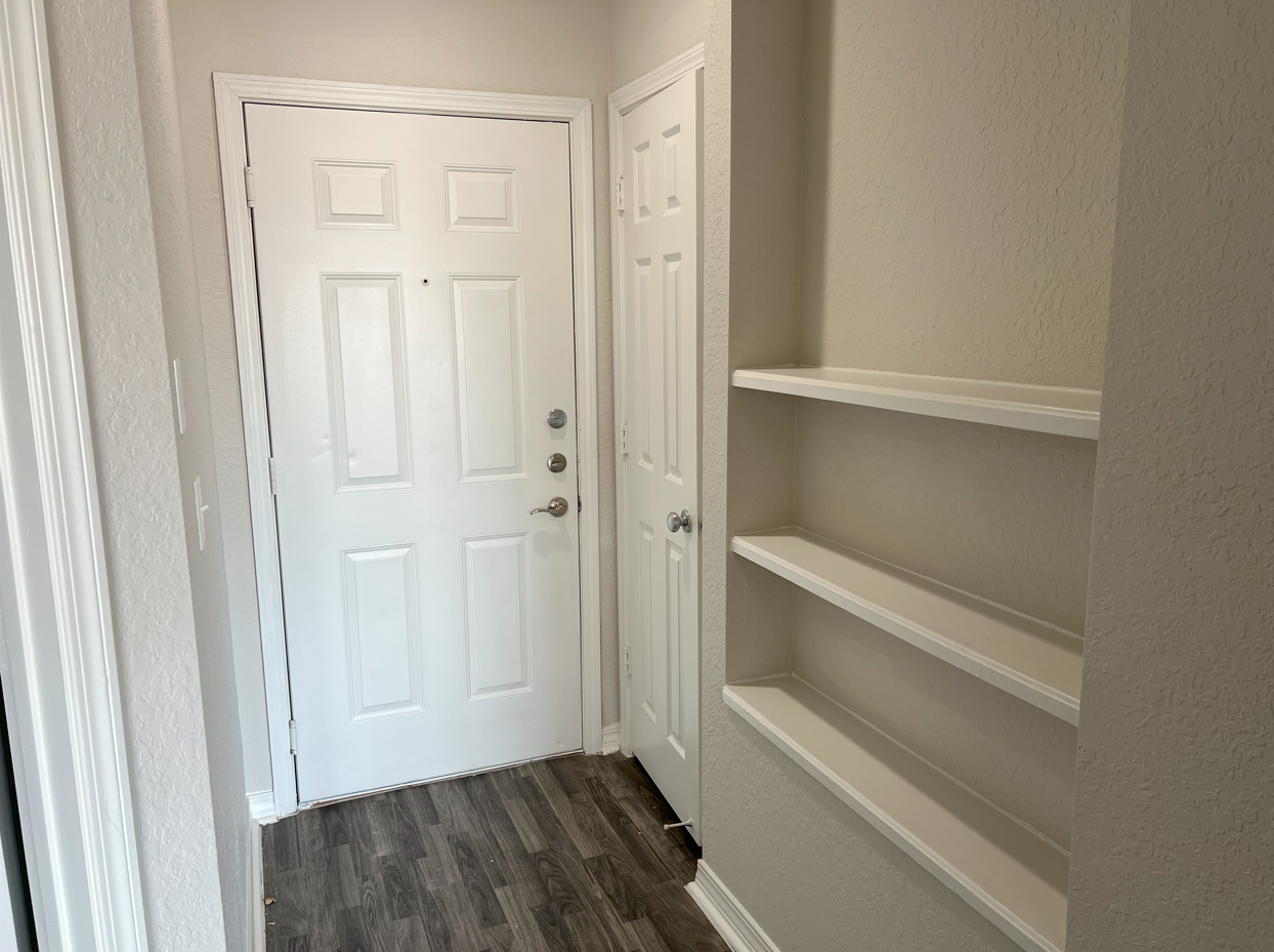 Narrow entry hallway with a white front door, gray wood floor, and built-in wall shelves.