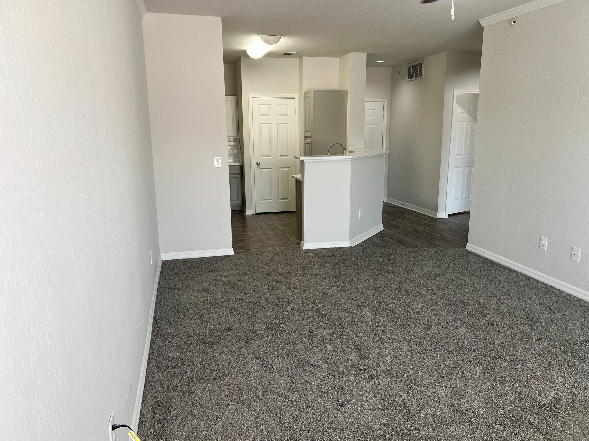 Empty carpeted apartment living room with white walls, ceiling fan, and kitchen pass-through counter
