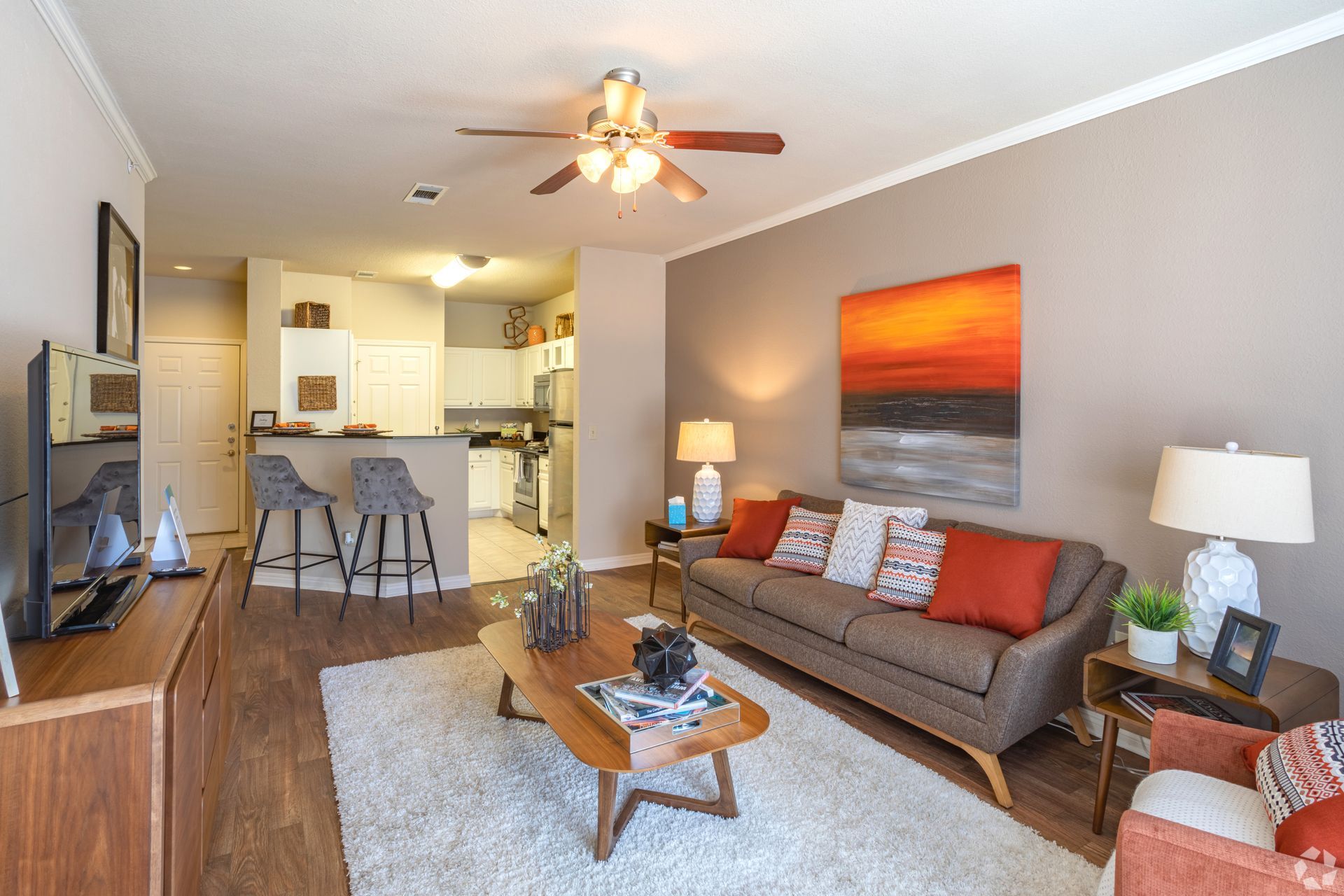 Bright living room with gray sofa, orange wall art, coffee table, and open kitchen with bar stools