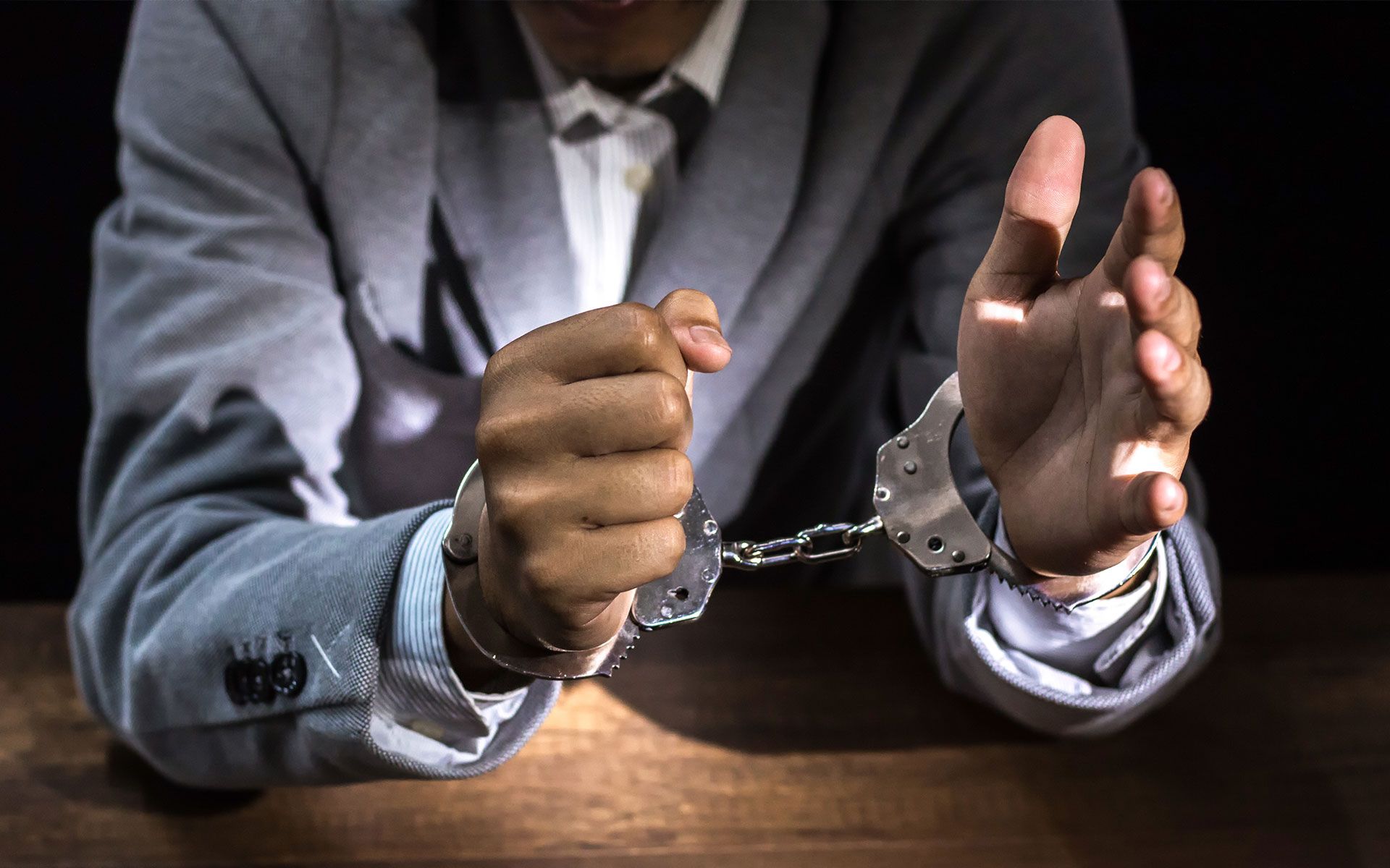 A man in a suit is sitting at a table with his hands in handcuffs.