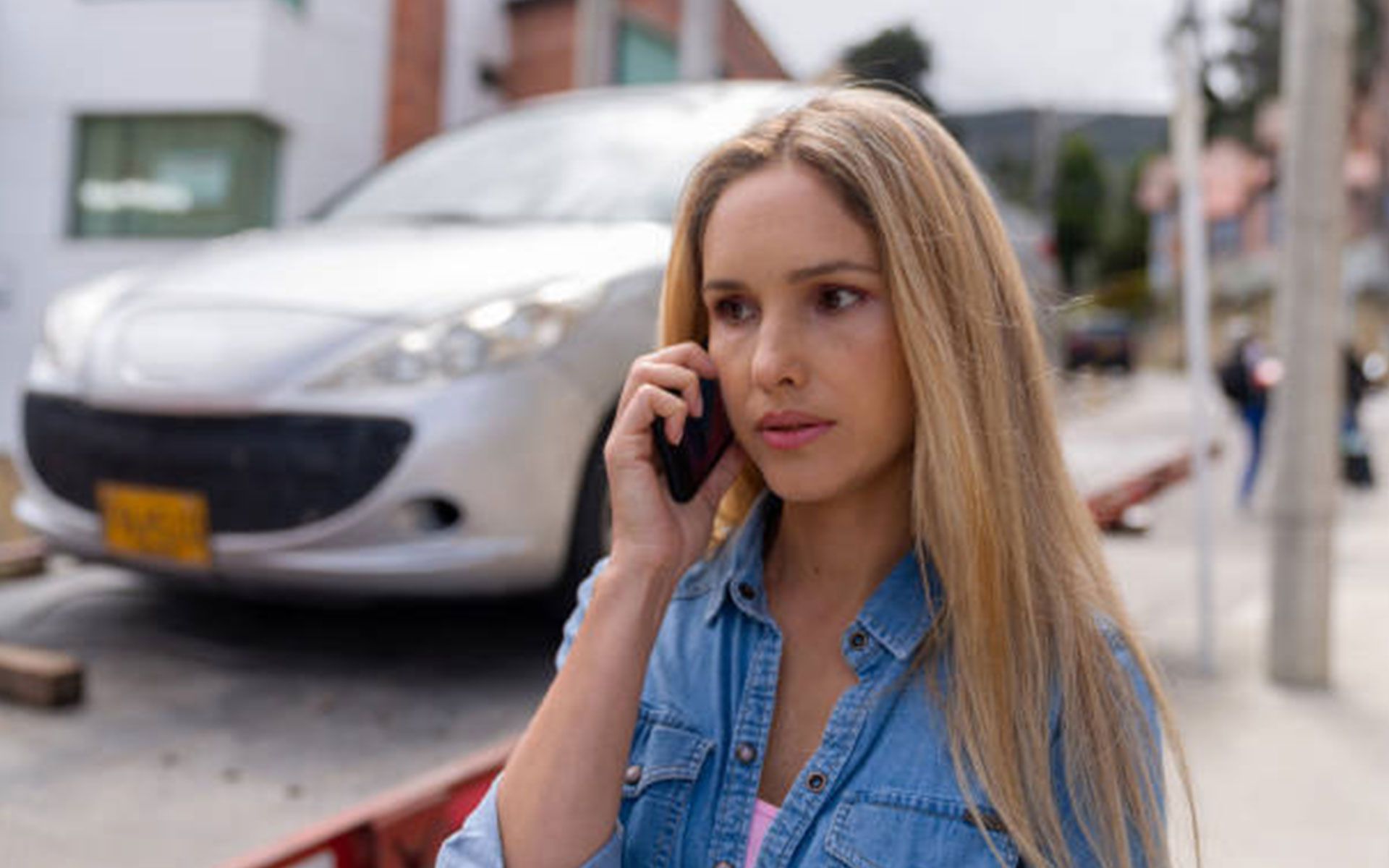 A woman is talking on a cell phone in front of a white car.