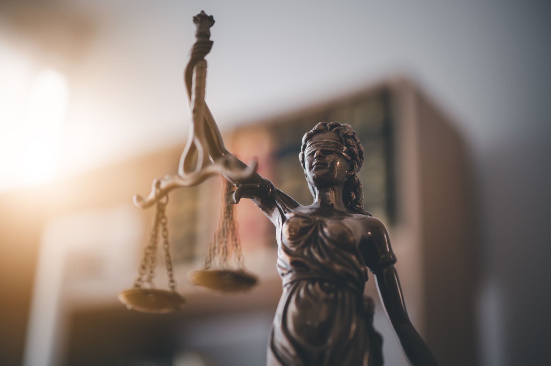 Bronze Lady Justice statue holding scales, with a blurred courtroom in the background