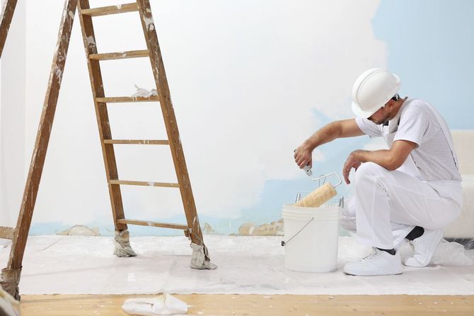 Un imbianchino in tuta da lavoro bianca dipinge un muro accanto a una scala, con un secchio di vernice sul pavimento.
