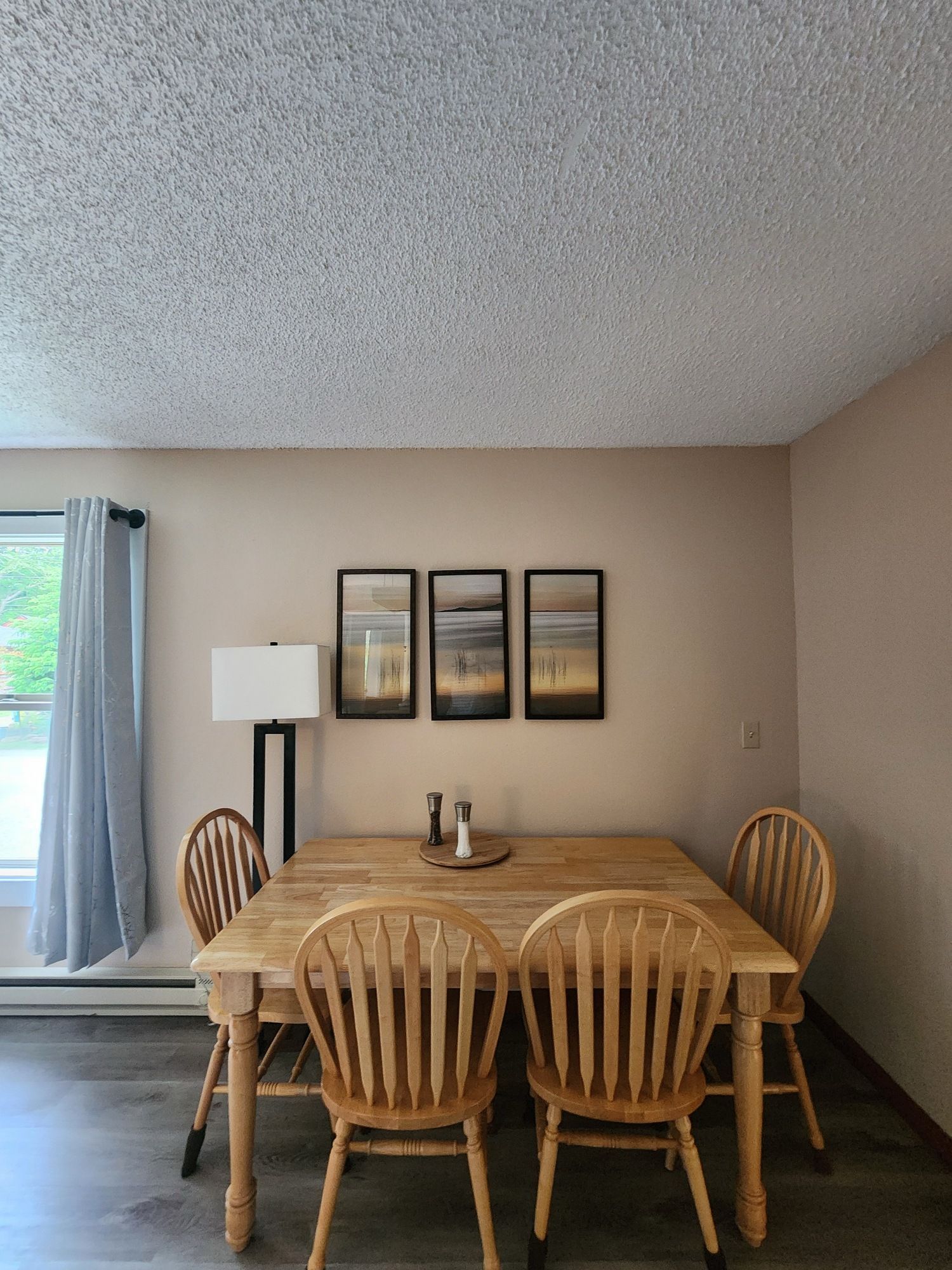 Dining area with a wooden table, six chairs, floor lamp, and framed wall art beside a window.