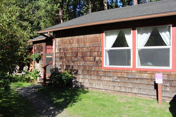 Small brown shingled house with red-trimmed windows and a grassy yard in daylight