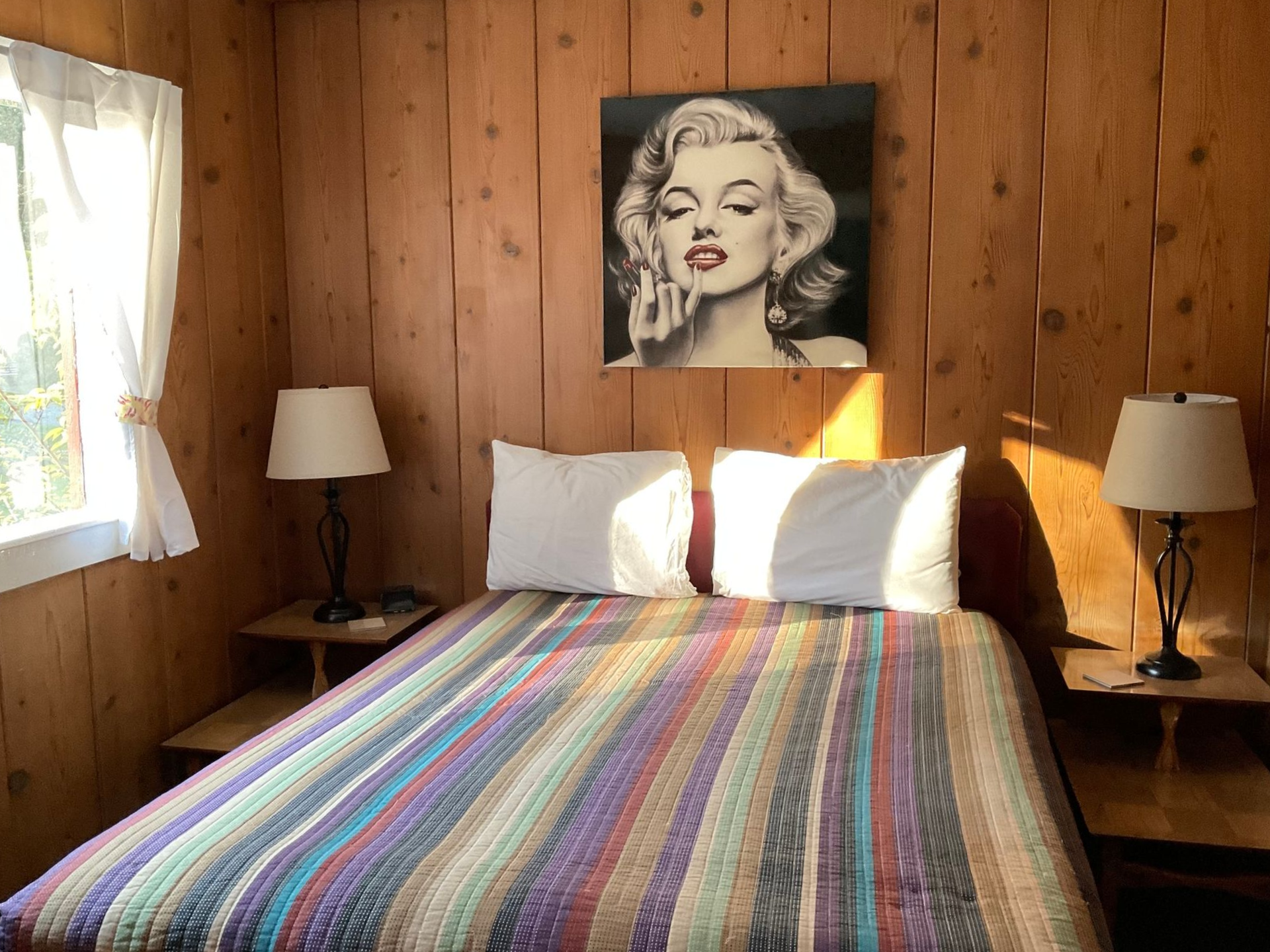 Wood-paneled bedroom with a striped bedspread, two lamps, and a Marilyn Monroe portrait above the bed