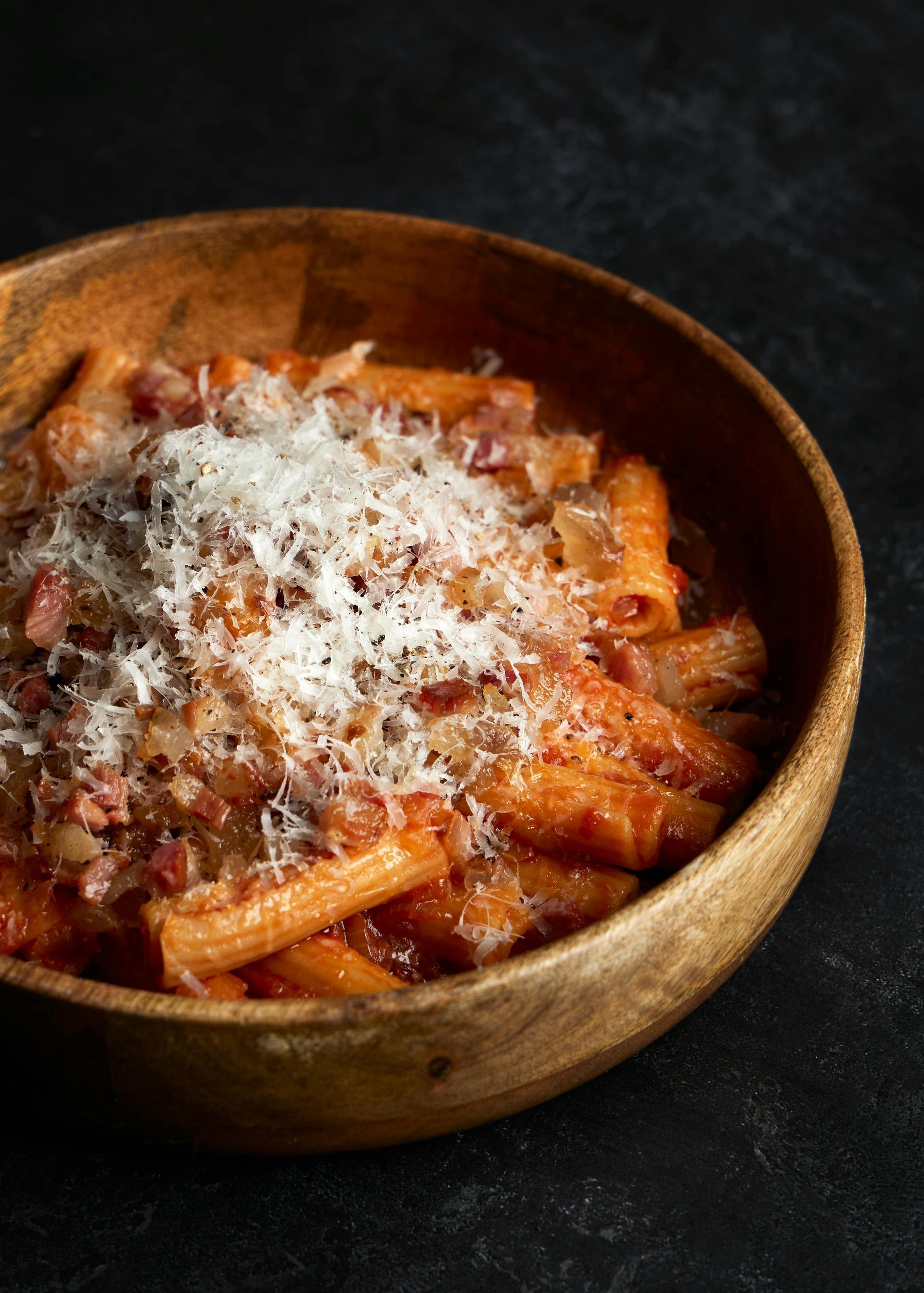 Wooden bowl of penne pasta with tomato sauce and grated cheese on a dark surface