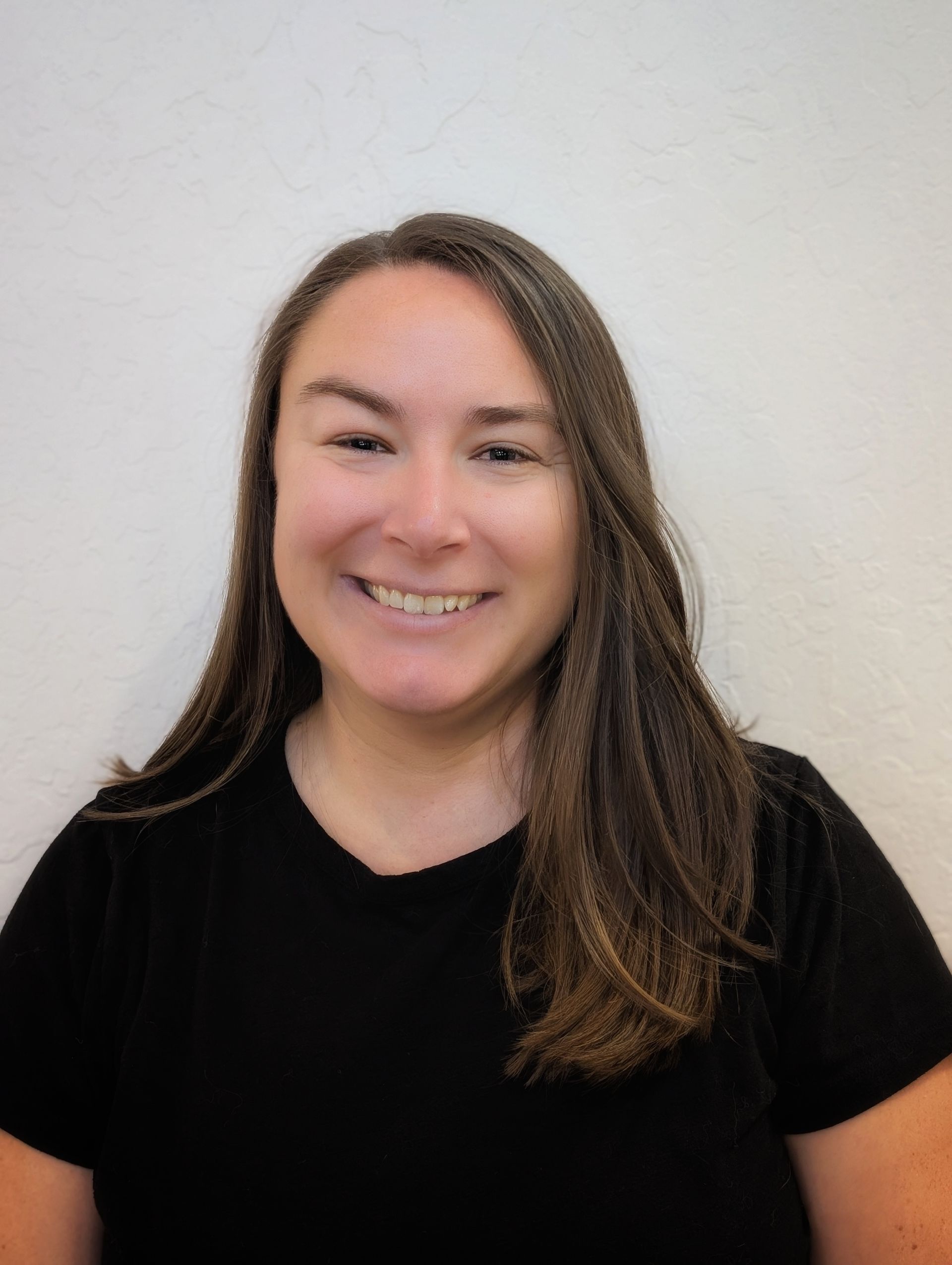 Woman with long brown hair smiles, wearing a black shirt against a plain white wall.