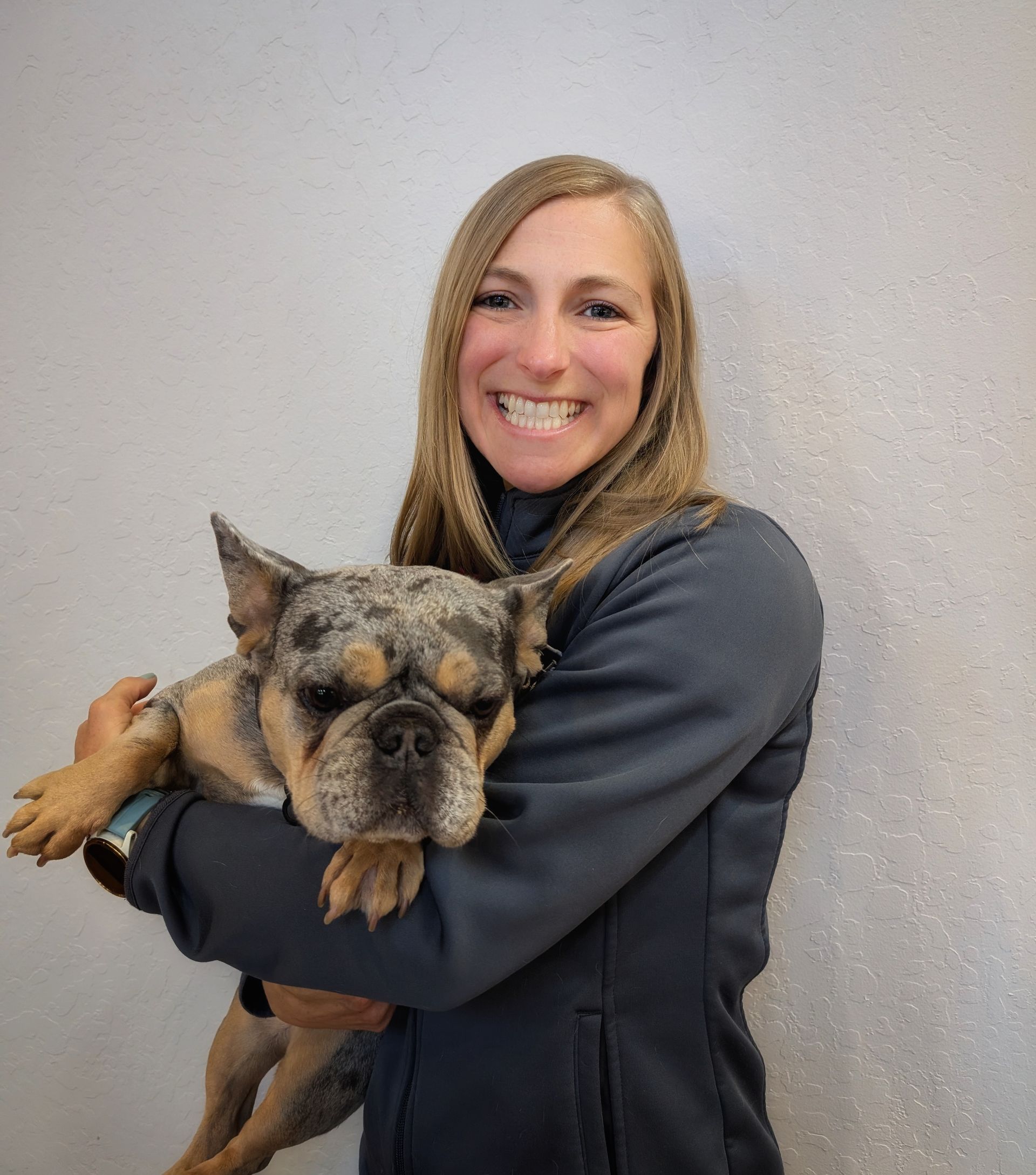 Woman in gray jacket holds dog with mottled fur, smiling against a light gray wall.