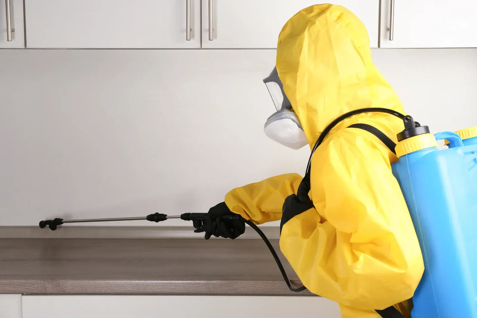 Man in protective gear sprays pesticide near a kitchen cabinet and sink.