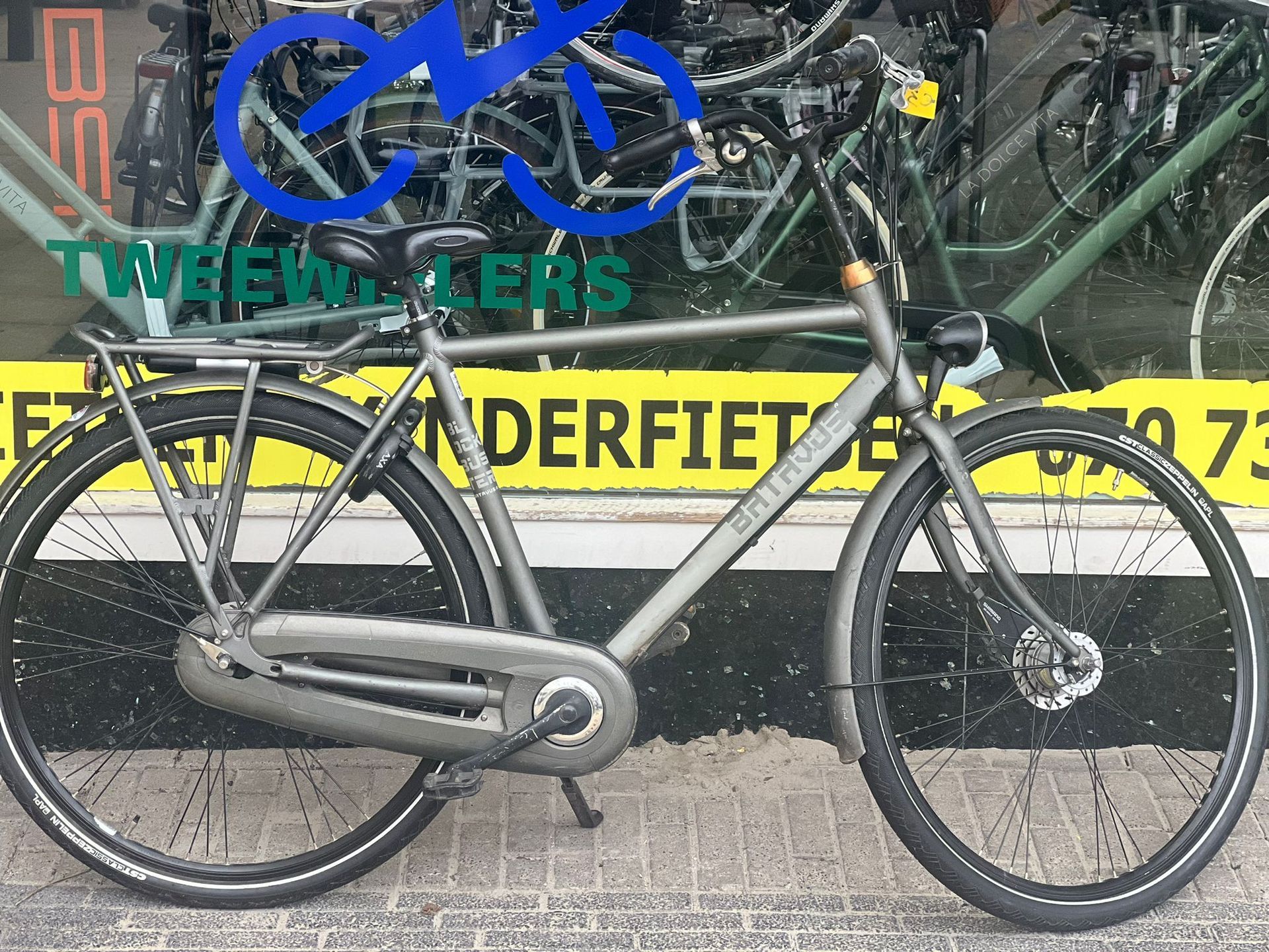 Grijze fiets in Hollandse stijl geparkeerd voor een winkel.