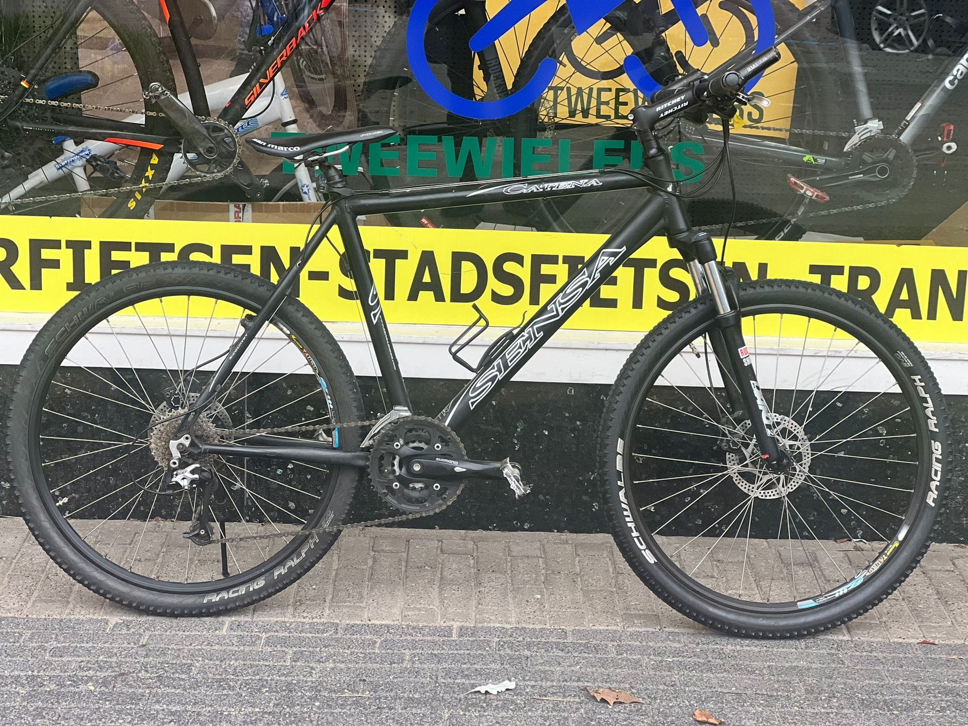 Een zwarte mountainbike staat geparkeerd voor de etalage van een fietsenwinkel, met schijfremmen en witte velgranden.
