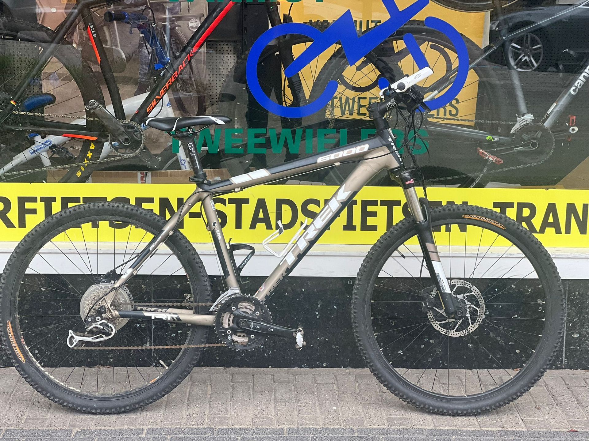 Een grijze en zwarte Trek mountainbike staat geparkeerd voor een fietsenwinkel, met het logo en de tekst van de winkel zichtbaar in het raam.