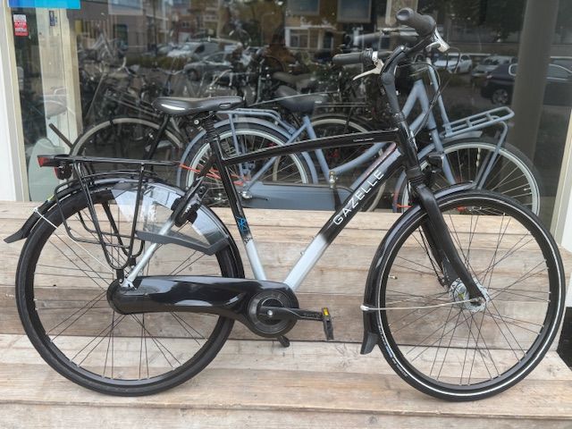Zwarte en zilverkleurige stadsfiets met bagagedrager, spatborden en witte banden, geparkeerd voor een winkel.