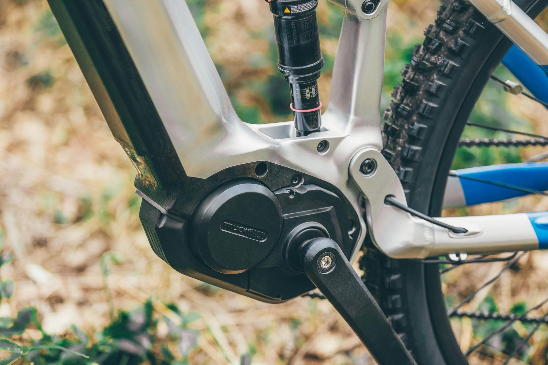 E-bike motor en crankstel, zilver en zwart, achterwiel zichtbaar, geplaatst in een natuurlijke buitenomgeving.