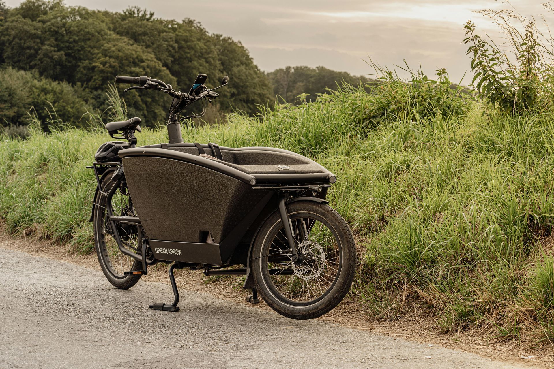 Zwarte elektrische bakfiets geparkeerd op een grindpad naast een grasheuvel; bomen op de achtergrond, bewolkte lucht.