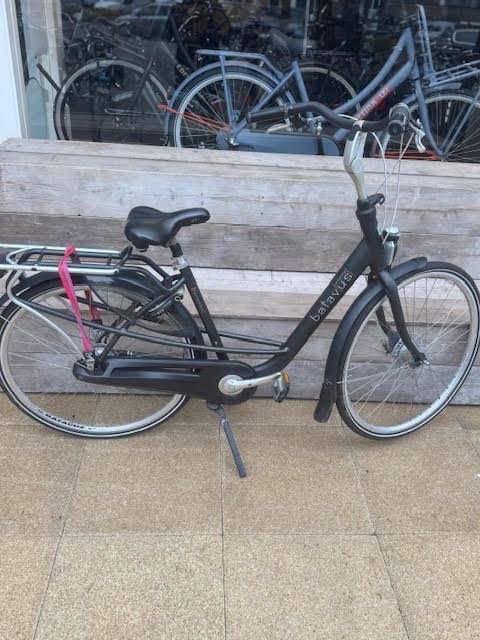 Een zwarte Gazelle-fiets staat buiten geparkeerd, voorzien van spatborden, een bagagedrager en een standaard.