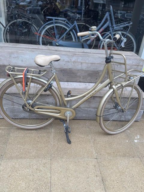 Gouden fiets met mandjes en crèmekleurig zadel, geparkeerd voor een winkelraam.