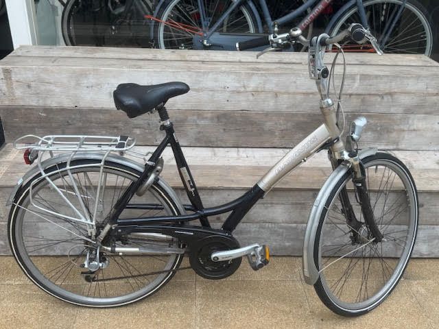 Zwarte en zilverkleurige fiets buiten geparkeerd, met spatborden voor en achter, bagagedrager en koplamp.