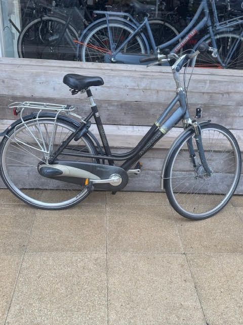 Een donkergrijze stadsfiets staat geparkeerd voor een winkelraam; op de achtergrond zijn andere fietsen zichtbaar.