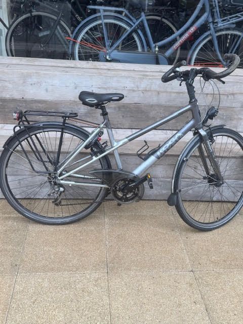 Zilverkleurige hybride fiets met zwarte accenten, spatborden voor en achter en een bagagedrager, staat buiten geparkeerd.